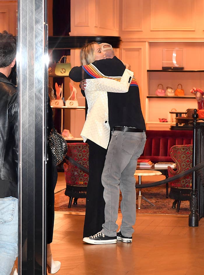 Tish Cyrus and Dominic Purcell hug in a high-end boutique with handbags displayed on shelves in the background