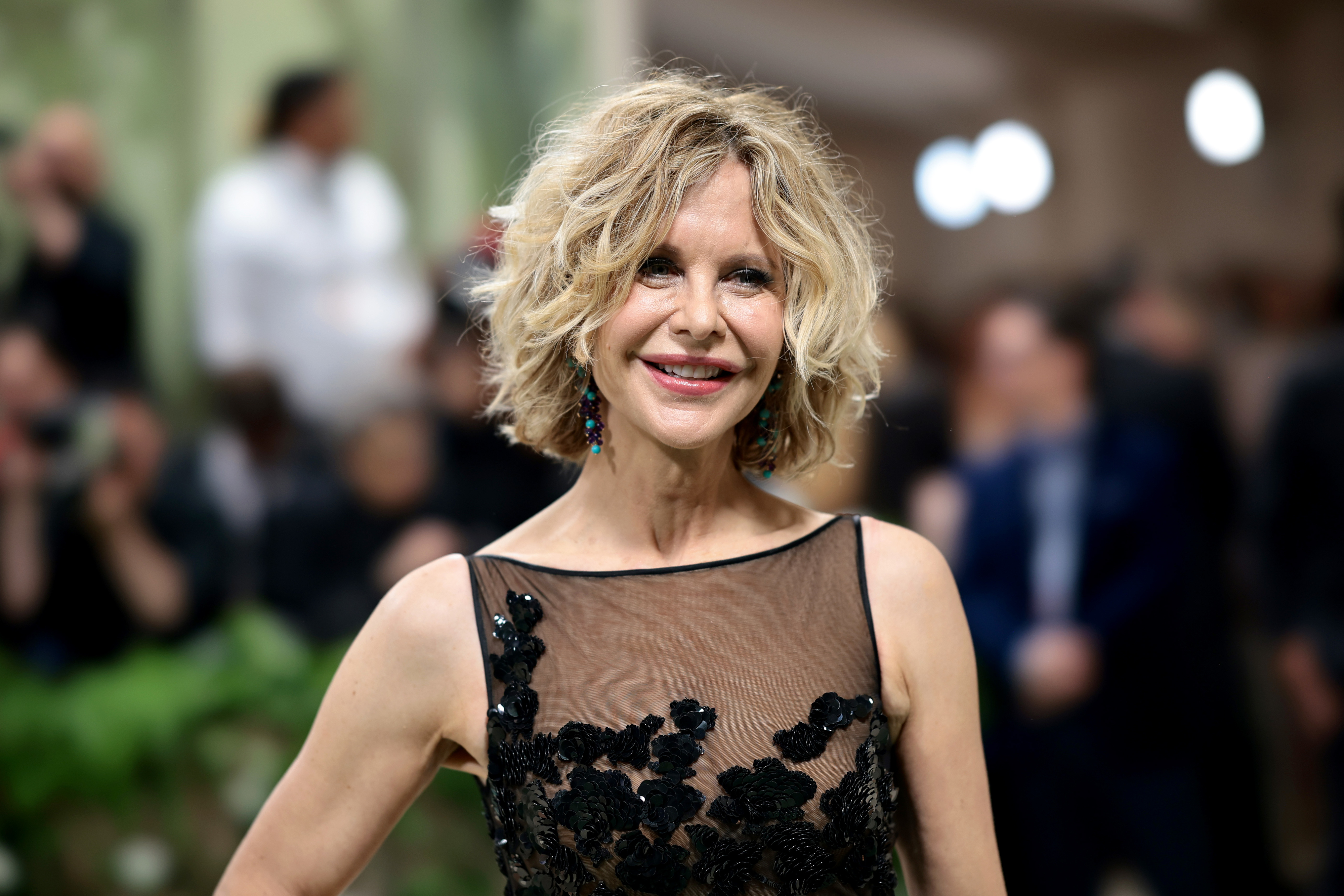 Meg Ryan smiling on the red carpet, wearing a sheer black dress with floral detailing