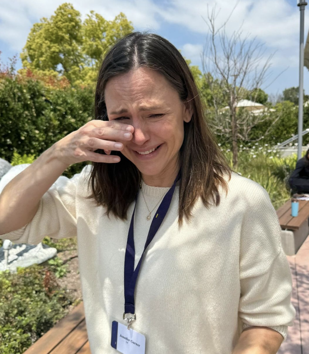 Jennifer Garner standing outside, wiping tears from her eyes, wearing a cream sweater with a blue lanyard and name tag. Trees and a pathway are in the background