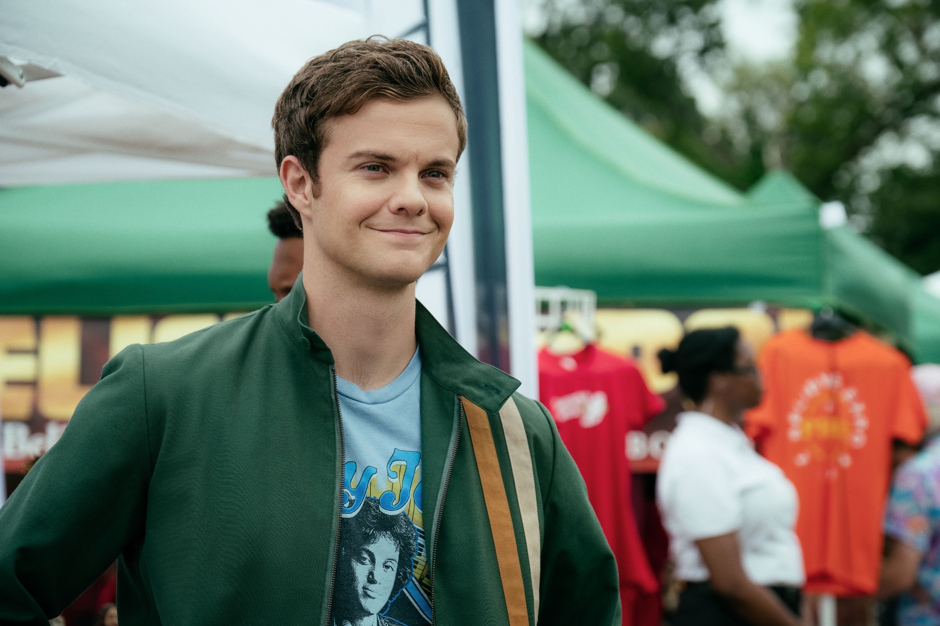 Jack Quaid at an outdoor event, wearing a casual jacket over a graphic T-shirt