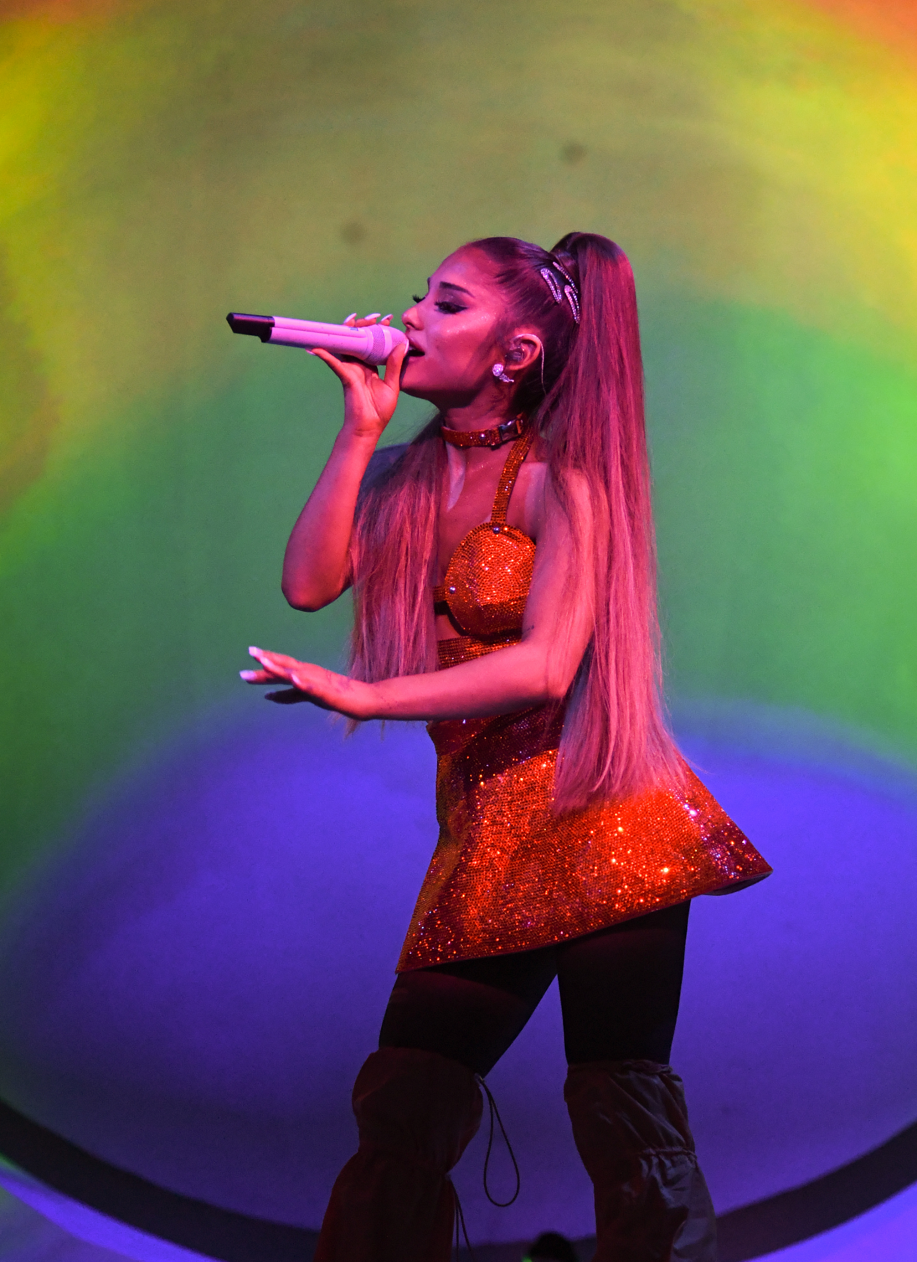Ariana Grande performing on stage, wearing a sparkly short dress and thigh-high boots, holding a microphone