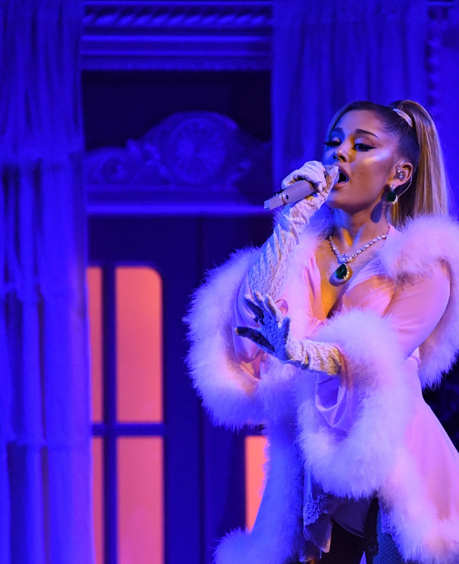 Ariana Grande performs on stage wearing a fur-trimmed outfit and gloves, singing into a microphone with her hair in a high ponytail