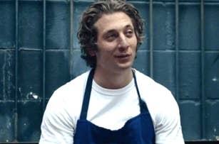 Jeremy Allen White in a white shirt and blue apron stands in front of a metal fence, likely a scene from a TV show or movie