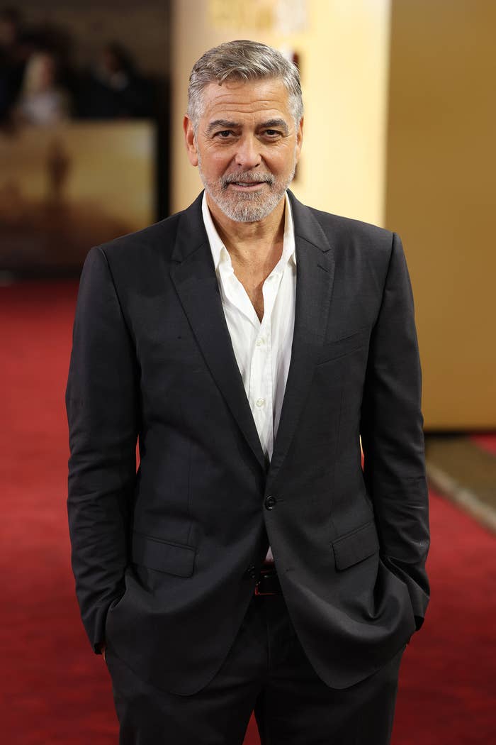 George Clooney stands on a red carpet, wearing a black suit with an open-collared white shirt