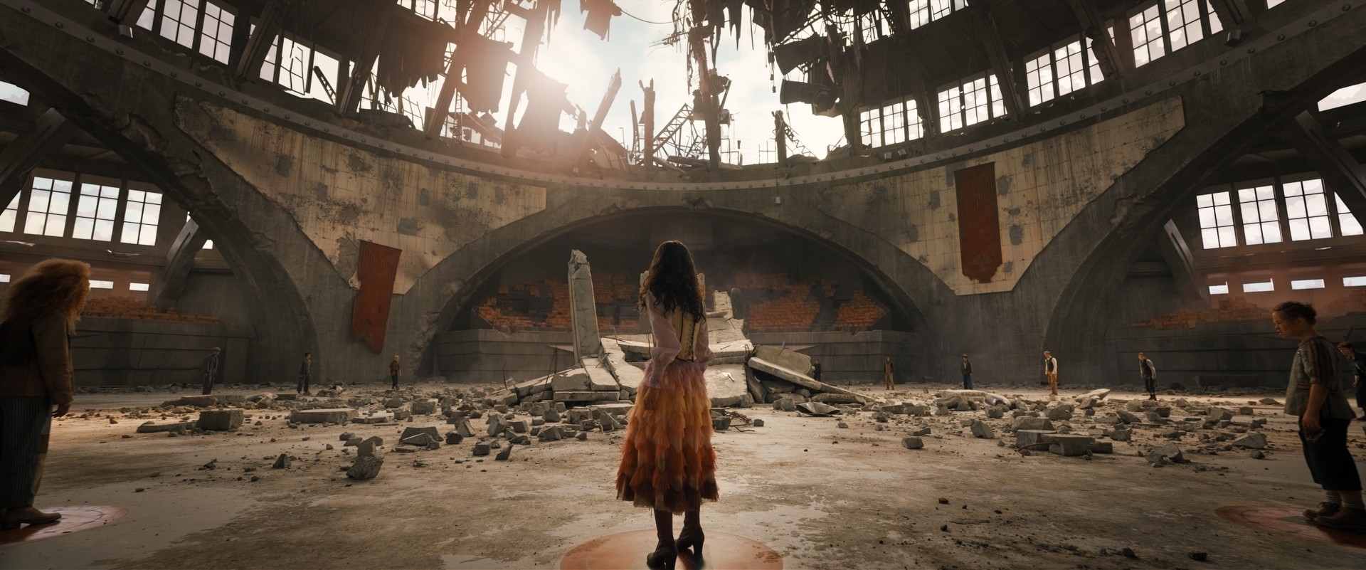 A woman stands in the middle of an abandoned, crumbling building with other people spread throughout the space, facing away from the camera