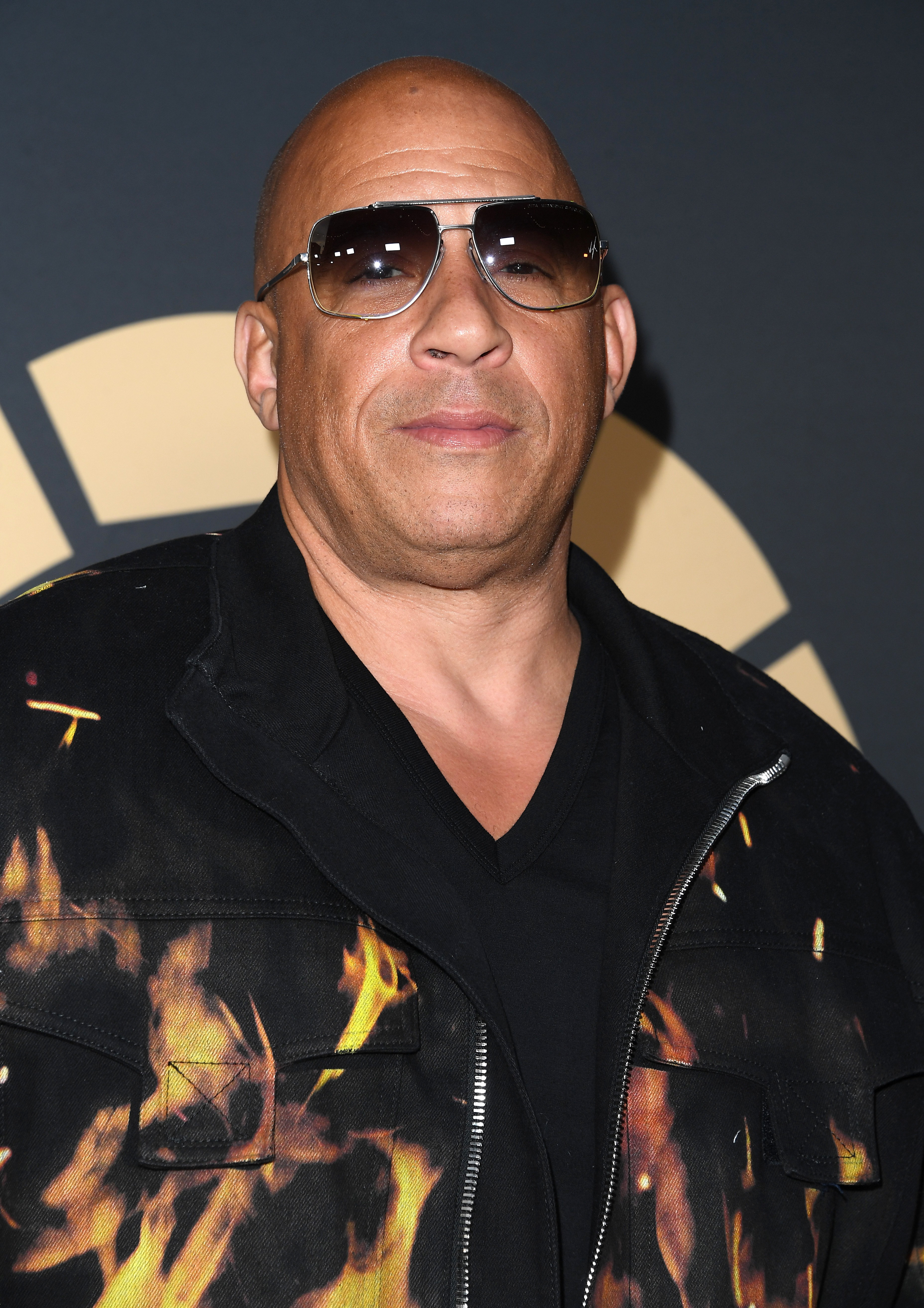 Vin Diesel wearing a black jacket with orange flame patterns and sunglasses, and standing in front of a promotional backdrop