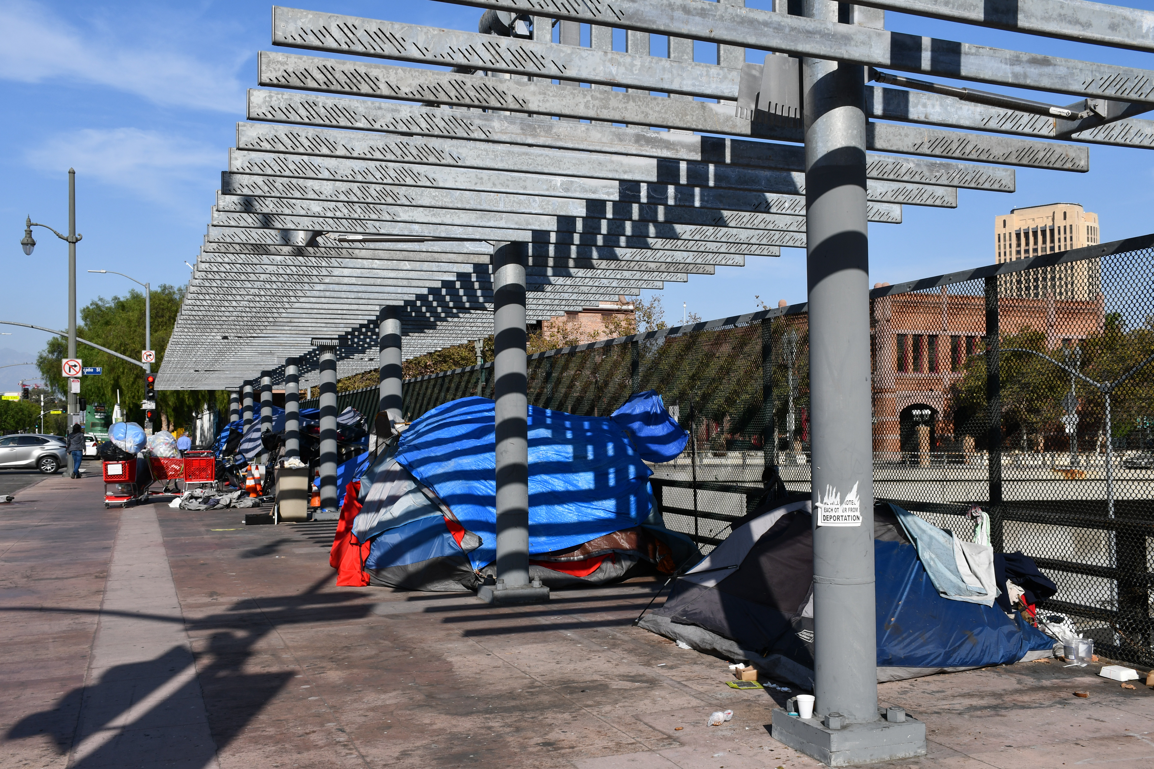 Row of makeshift tents and belongings under a shaded structure along a sidewalk, with buildings in the background