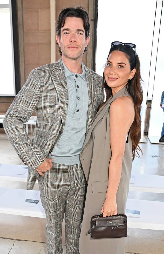 John Mulaney in a plaid suit and Olivia Munn in a sleeveless blazer with a handbag, standing together and posing for the camera