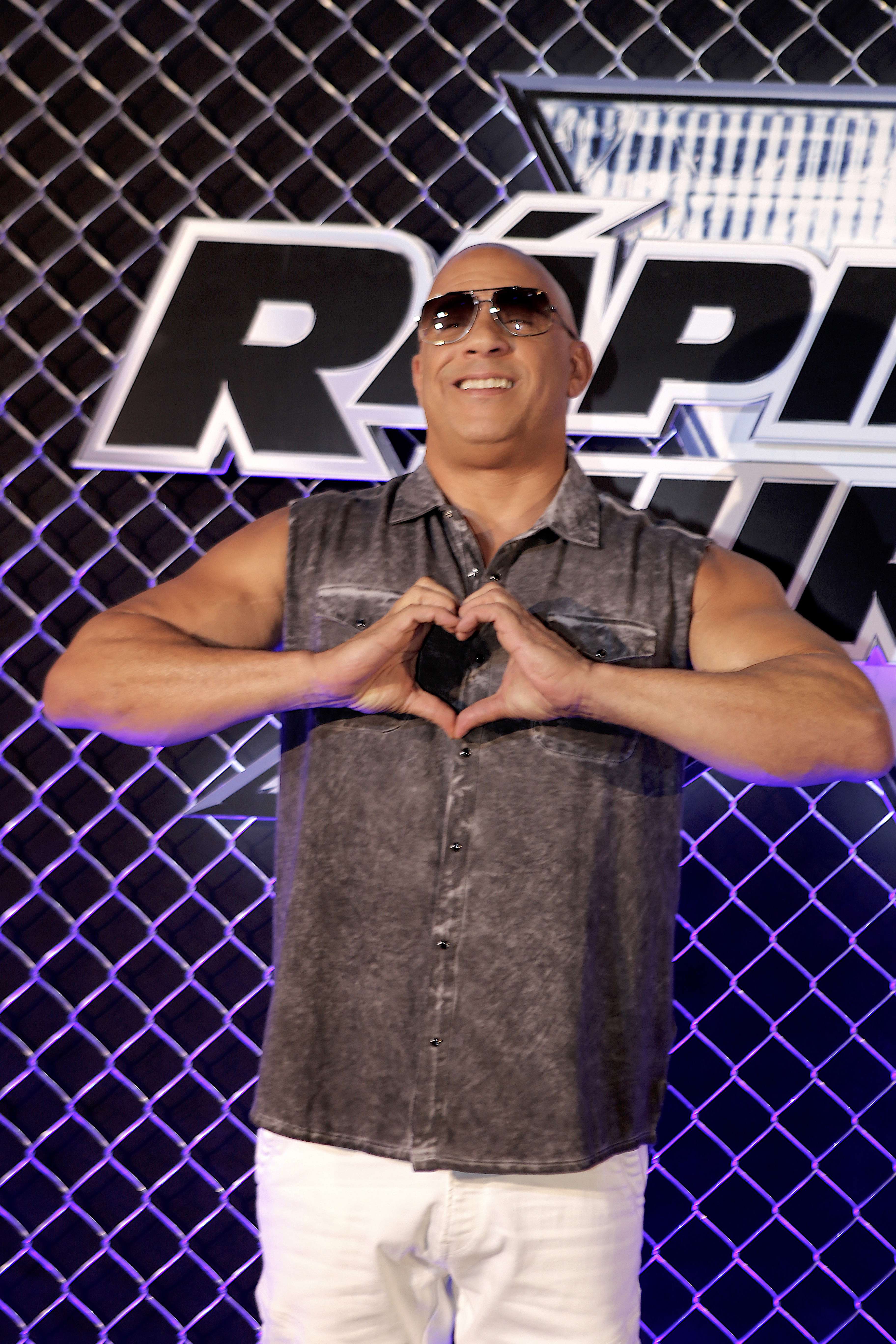 Vin Diesel stands in front of a backdrop, wearing sunglasses, a sleeveless denim shirt, and white pants, making a heart shape with his hands