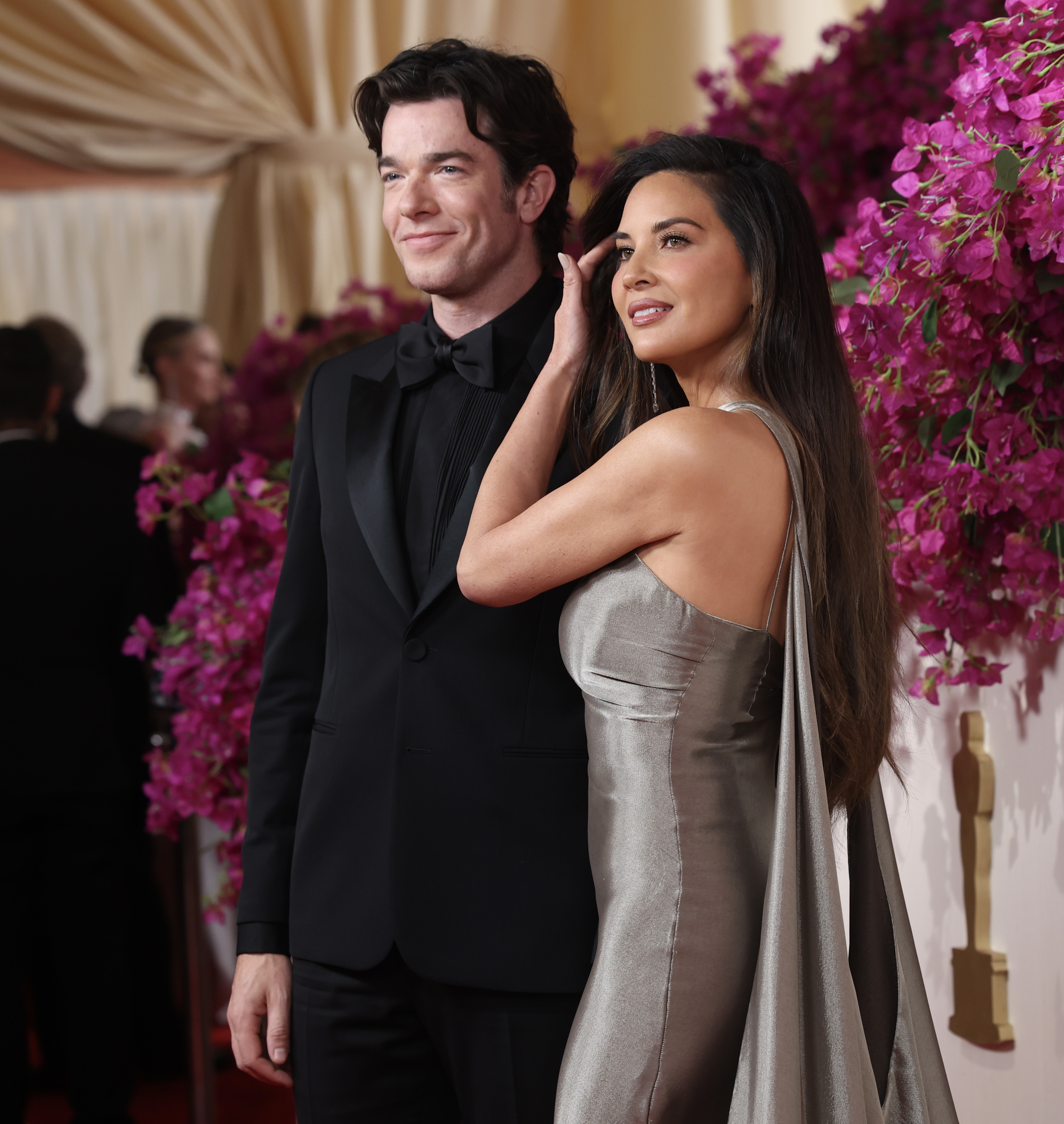 John Mulaney and Olivia Munn pose together on a red carpet event. John wears a classic black tuxedo, and Olivia wears a silver gown with a draped back
