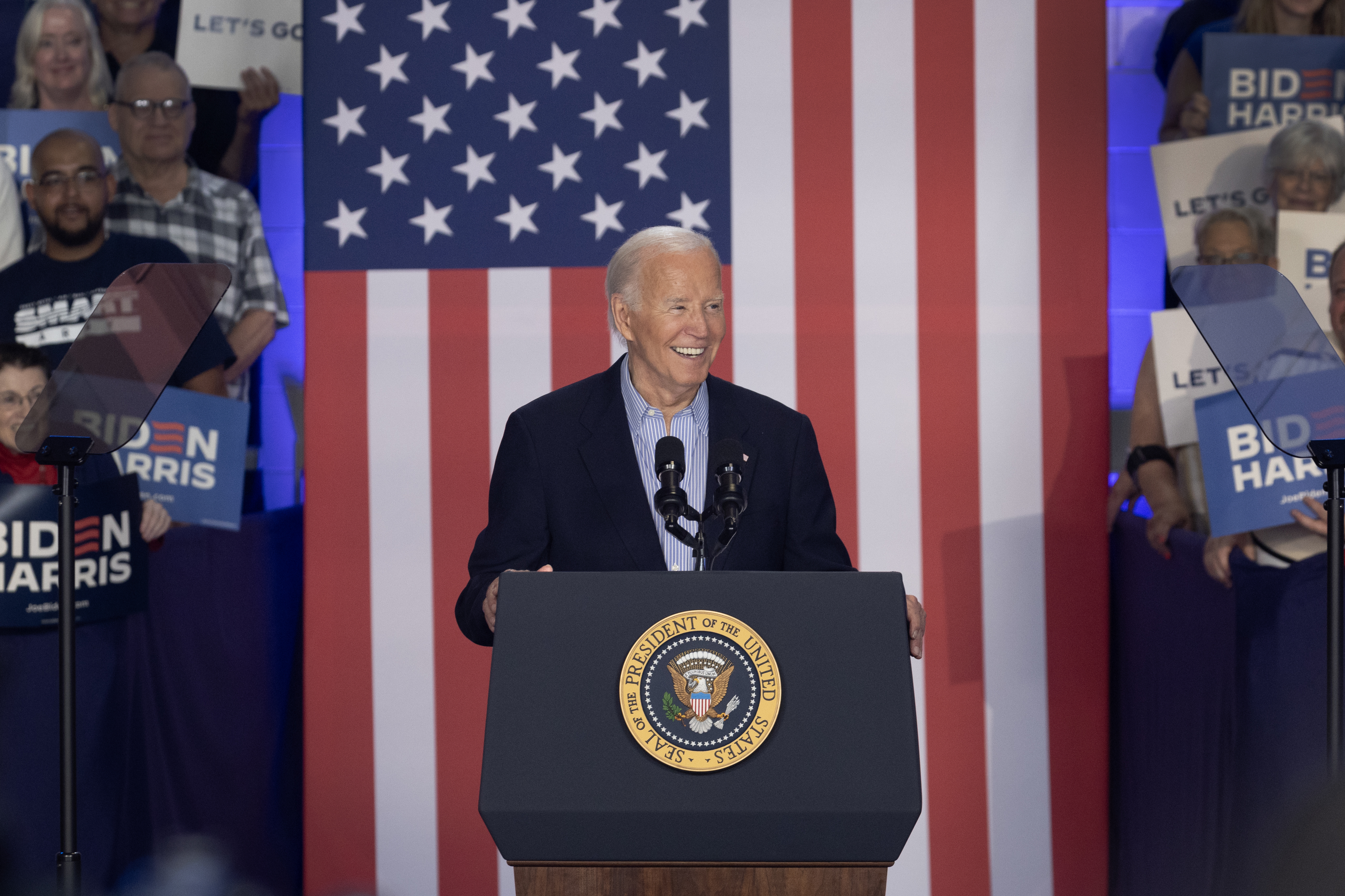 President Joe Biden speaking at a podium with an American flag backdrop. Audience members hold &quot;Biden-Harris&quot; and &quot;Let&#x27;s Go&quot; signs