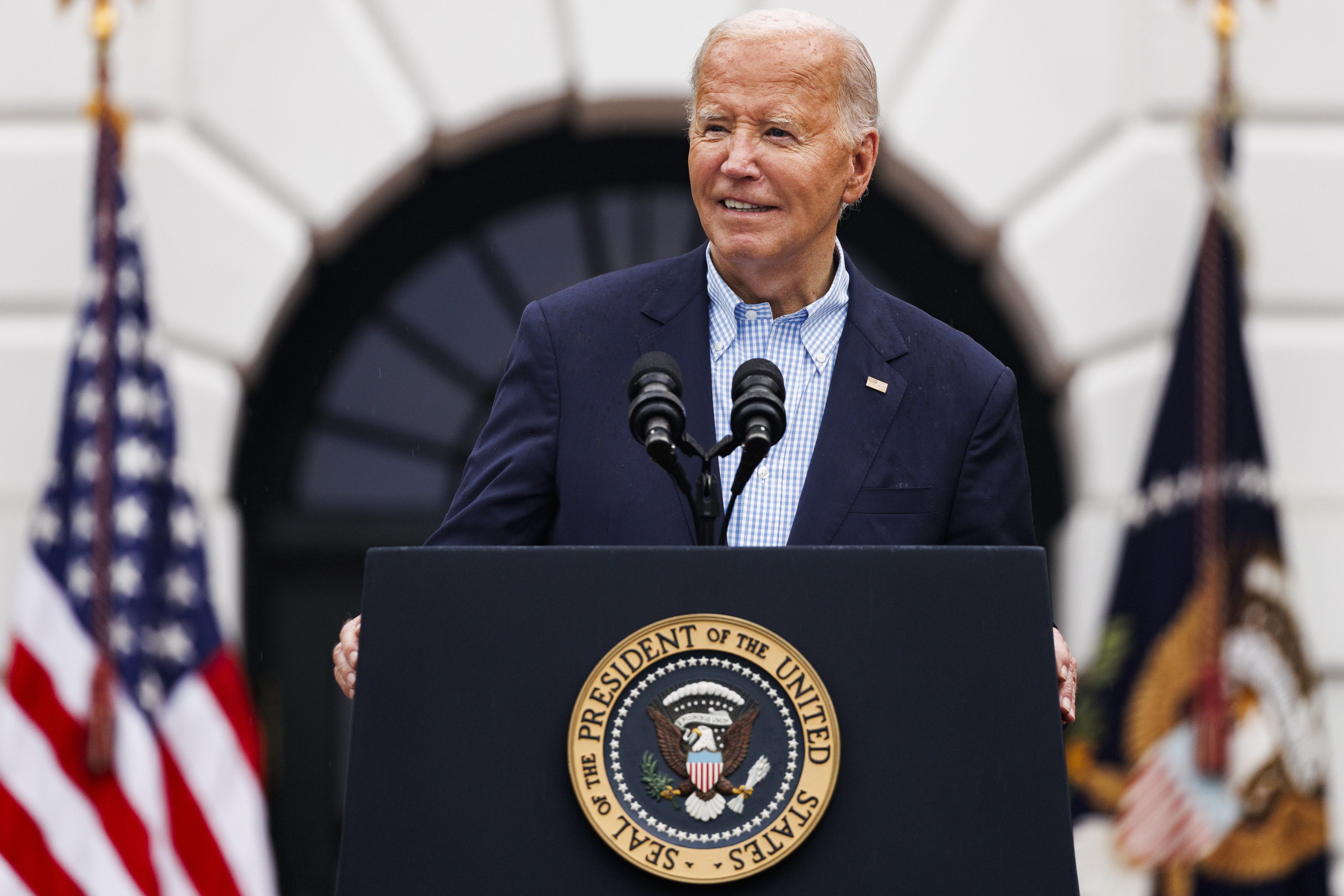 Joe Biden speaks at a podium with the presidential seal in front of microphones, American flags, and the White House backdrop