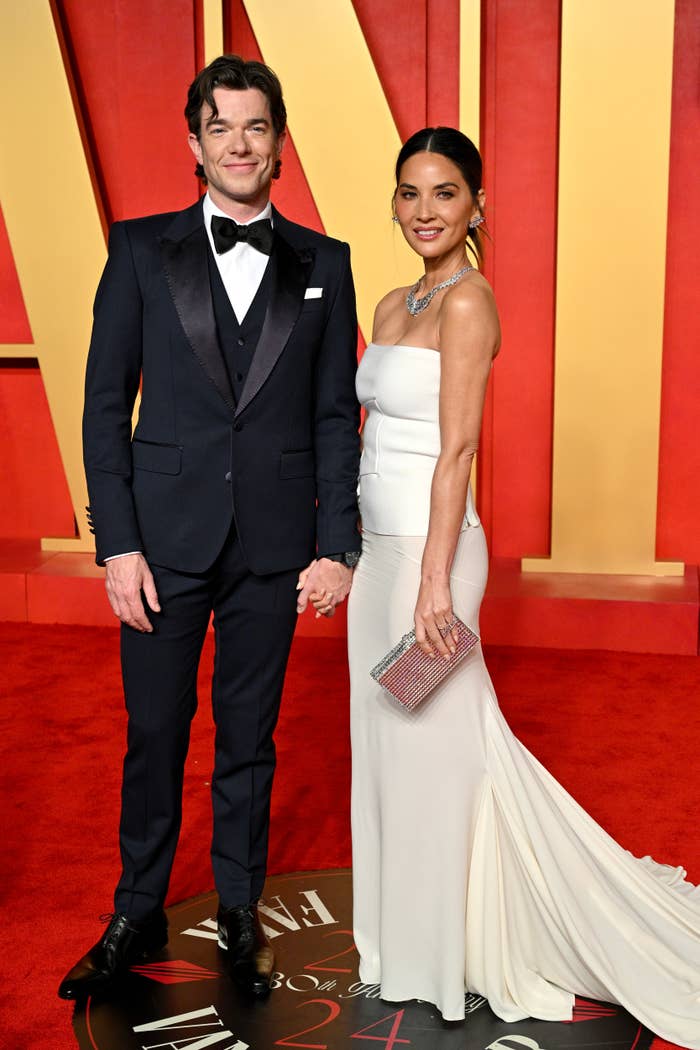 John Mulaney in a classic tuxedo and Olivia Munn in a strapless white gown, holding a sparkly clutch, are on the red carpet at a formal event