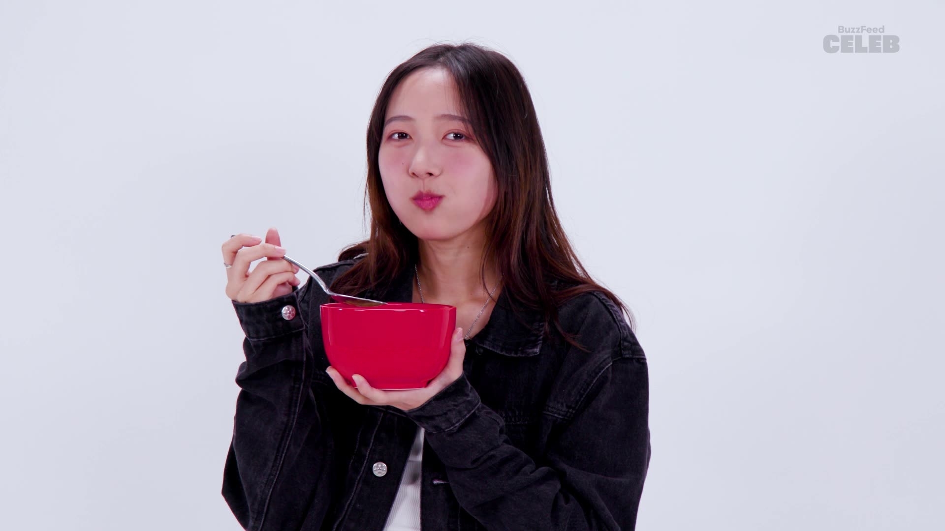 Hannah Bahng eats from a red bowl with a spoon while smiling. BuzzFeed Celeb logo in the corner