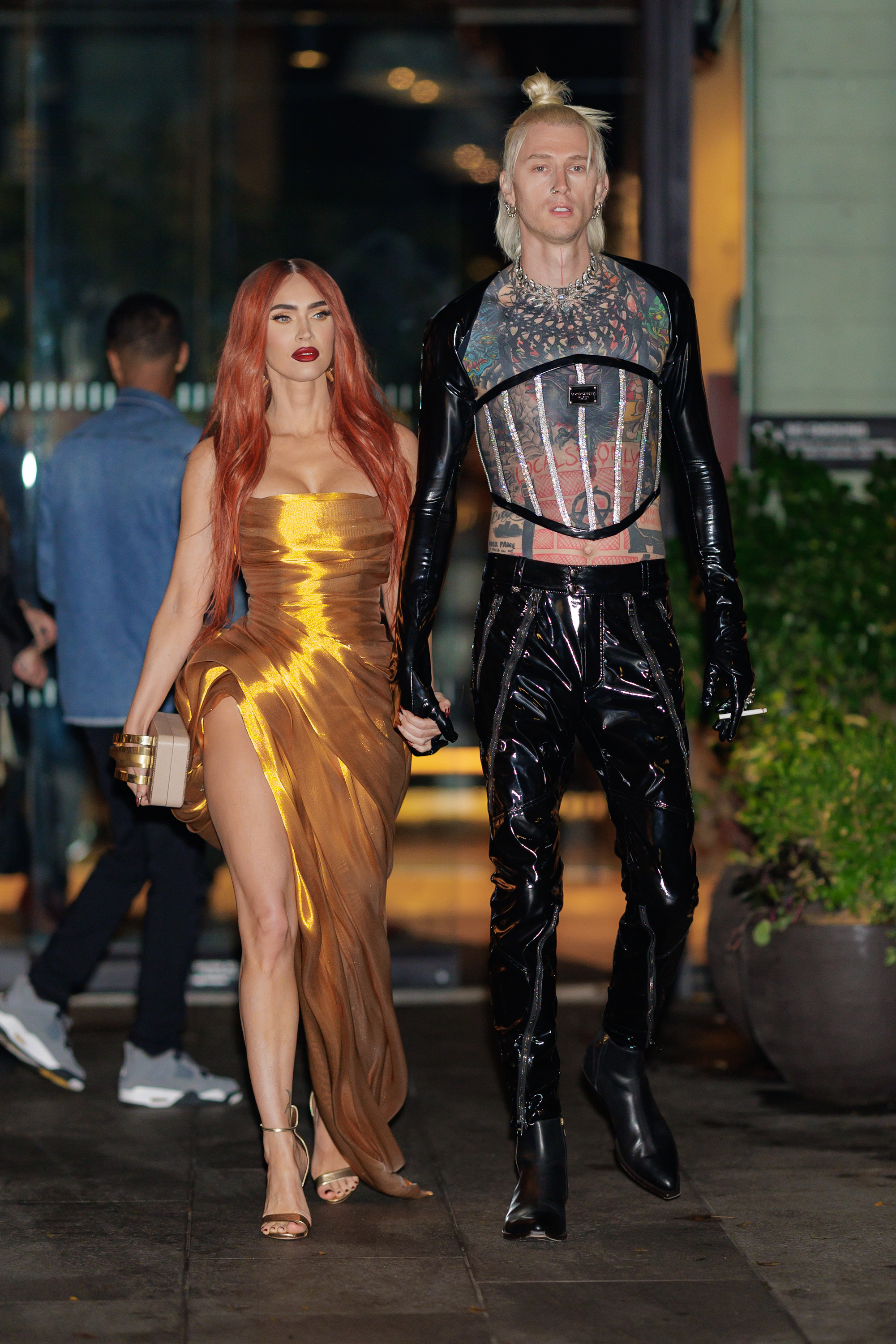Megan Fox in a strapless gown with high slit, holding hands with Machine Gun Kelly in a printed shirt, chest armor, and vinyl pants. Both walking outdoors