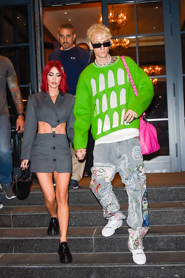 Megan Fox and Machine Gun Kelly step out, holding hands. Megan wears a cropped blazer and mini skirt, while Machine Gun Kelly wears a bright green patterned sweater and ripped jeans