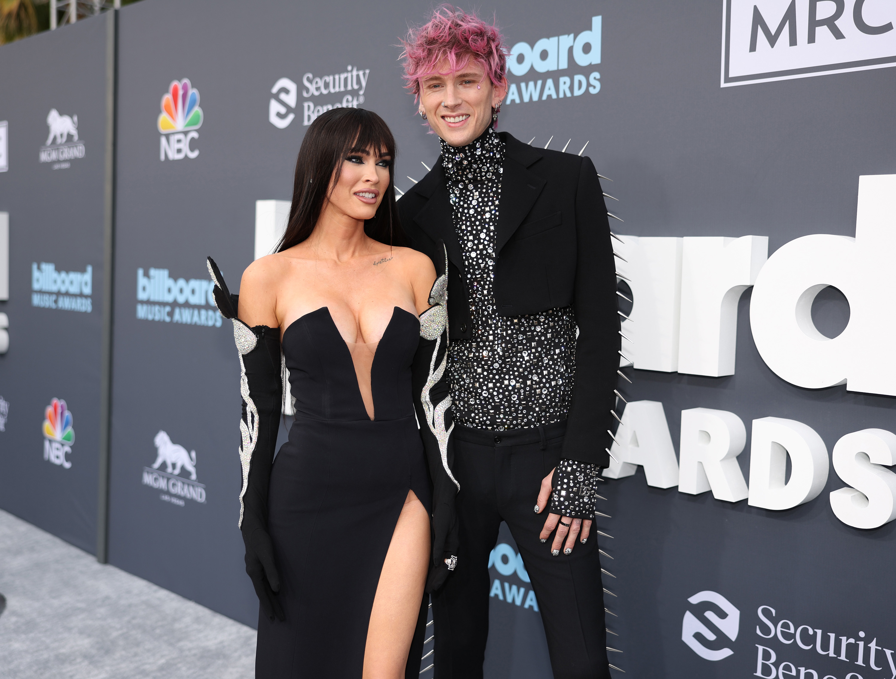 Megan Fox and Machine Gun Kelly pose on the red carpet at the Billboard Music Awards. Megan wears an off-shoulder dress with a high slit, and MGK wears a studded suit