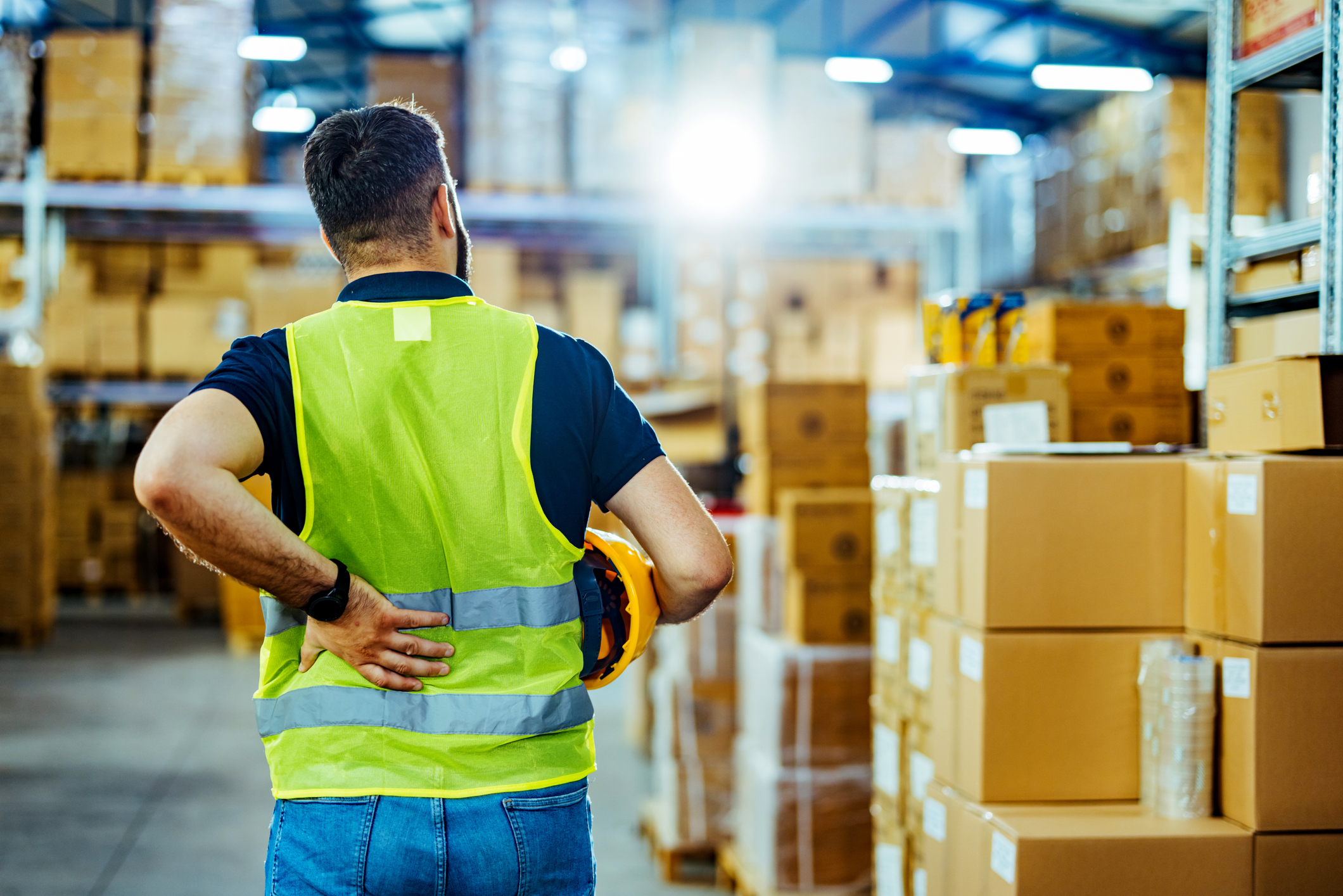 A person in a high-visibility vest stands in a warehouse holding their lower back, surrounded by shelves filled with cardboard boxes