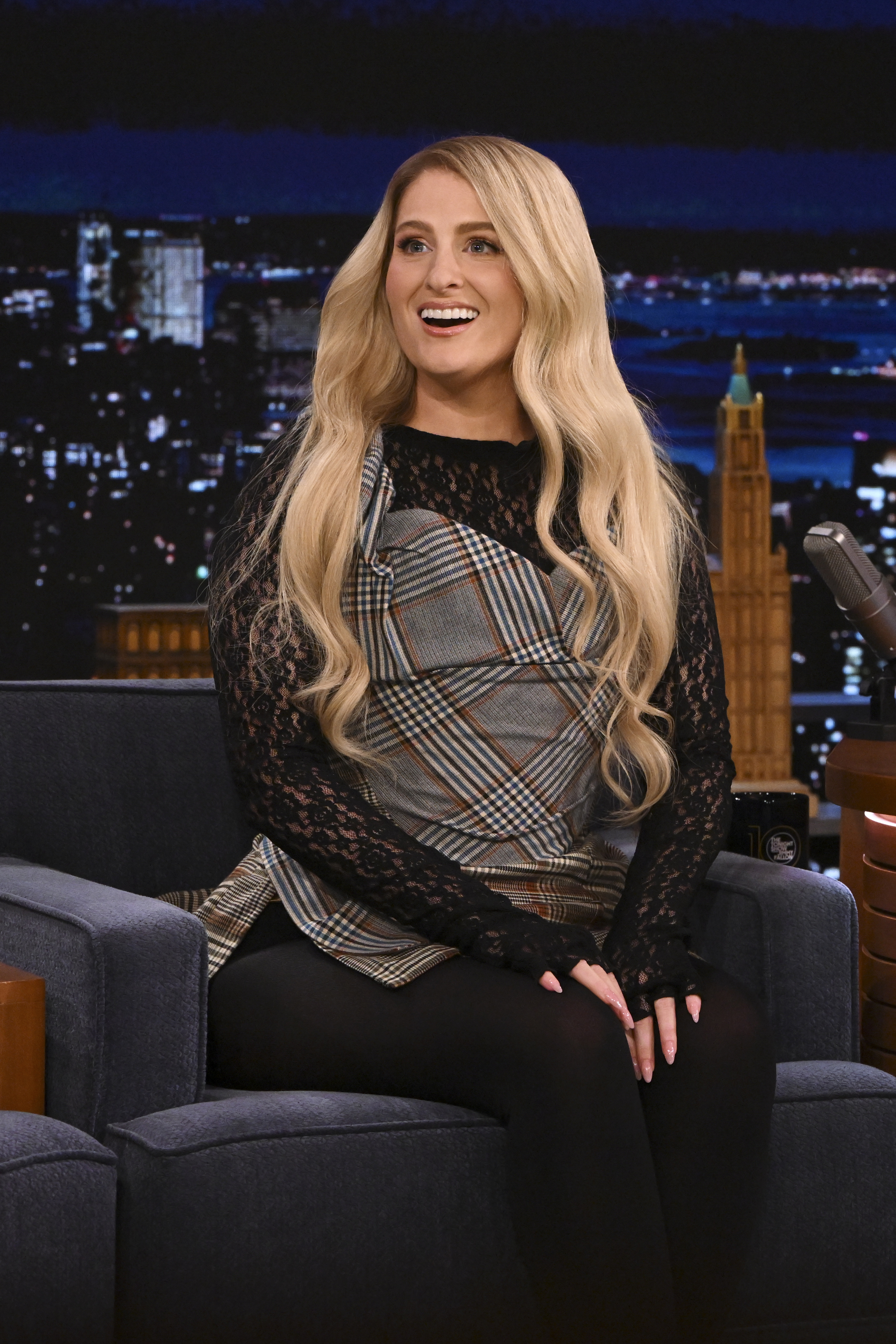 Meghan Trainor on a TV show set, wearing a patterned top with lace sleeves and leggings, seated and smiling