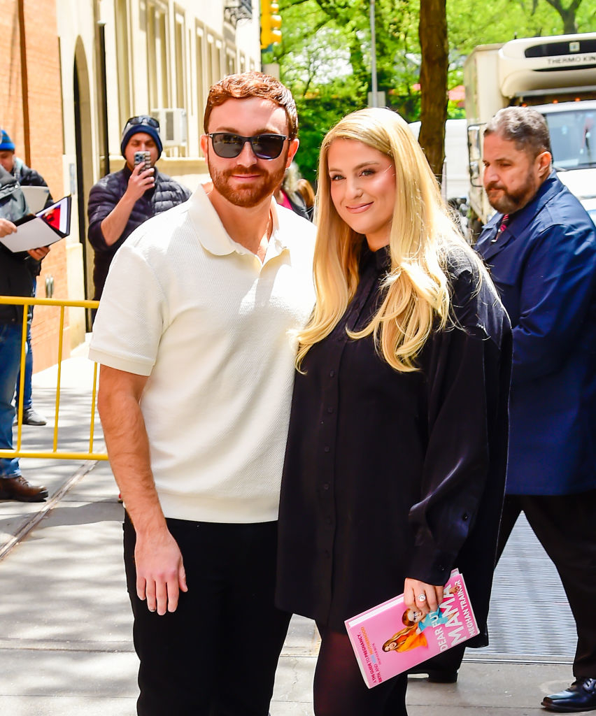 Meghan Trainor and Daryl Sabara pose together on a city sidewalk. Meghan wears a black dress shirt and holds a book, while Daryl wears a white polo and sunglasses