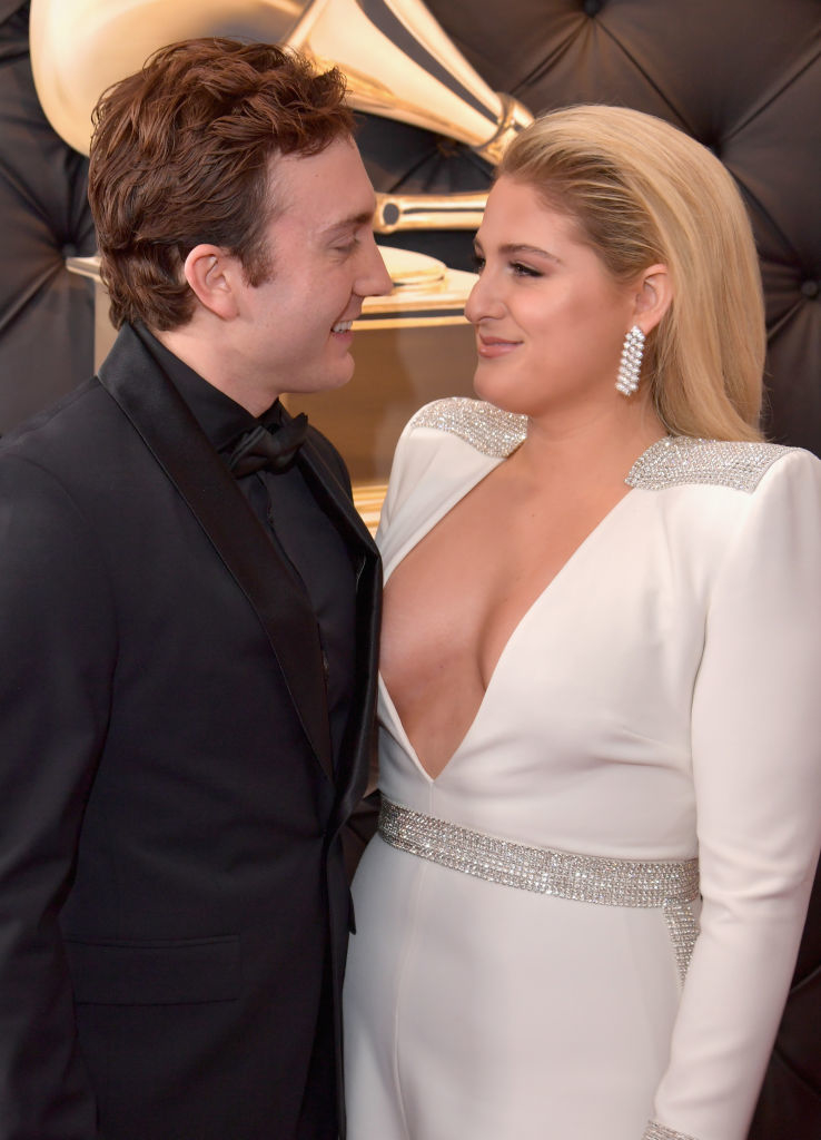 Meghan Trainor and Daryl Sabara pose together on a red carpet event. Meghan wears a deep V-neck gown with silver detailing, and Daryl wears a tuxedo