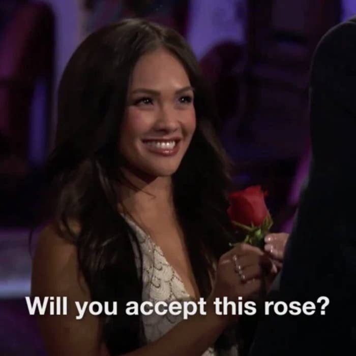 Jenn Tran from The Bachelor, smiles while offering a red rose to someone out of frame. Text: &quot;Will you accept this rose?&quot;