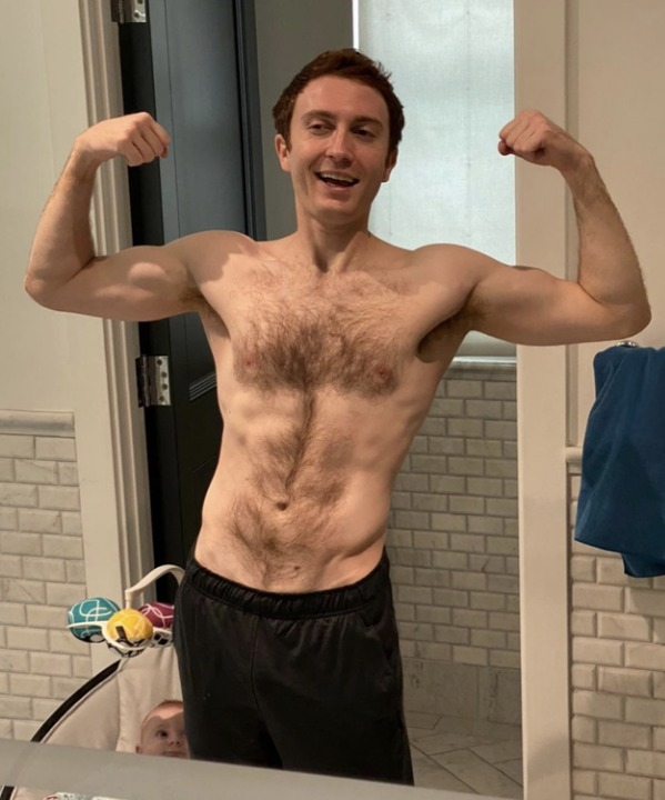 Daryl Sabara flexing muscles in front of a mirror, shirtless, wearing black shorts in a bathroom. Baby&#x27;s head visible at the bottom of the image. Names not provided