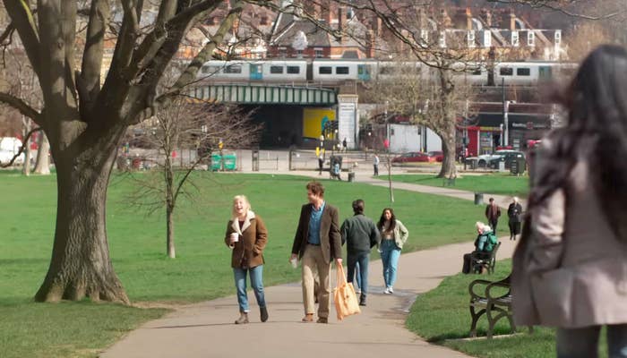 Andrew Garfield and Florence Pugh are walking in a park, with a train passing on a bridge in the background