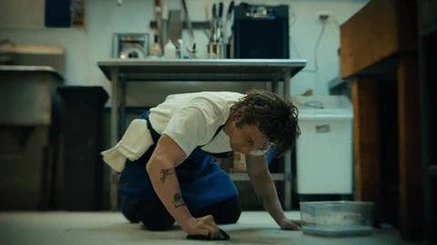 Jeremy Allen White, dressed in a white t-shirt and blue apron, scrubs the floor in a kitchen scene from a TV show