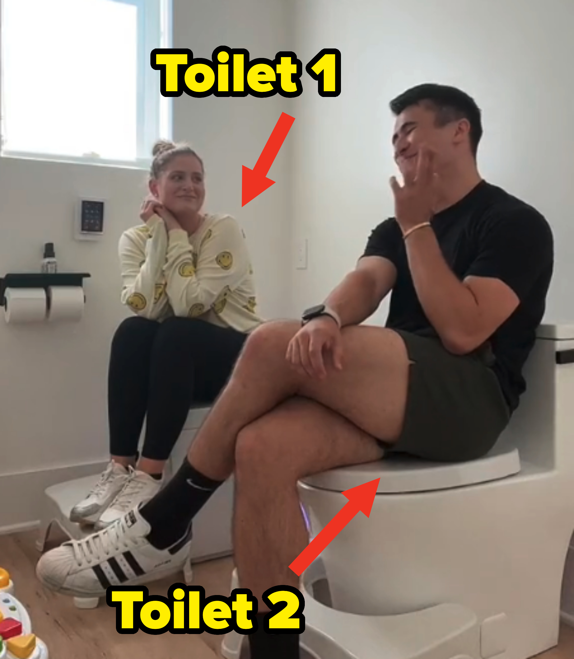 Meghan Trainor and a gentleman are sitting on adjacent toilets, smiling and engaged in conversation in a bathroom