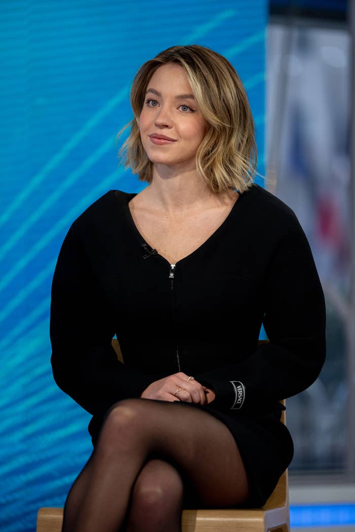 Sydney Sweeney is seated, wearing a dark outfit with sheer tights, on a talk show set with