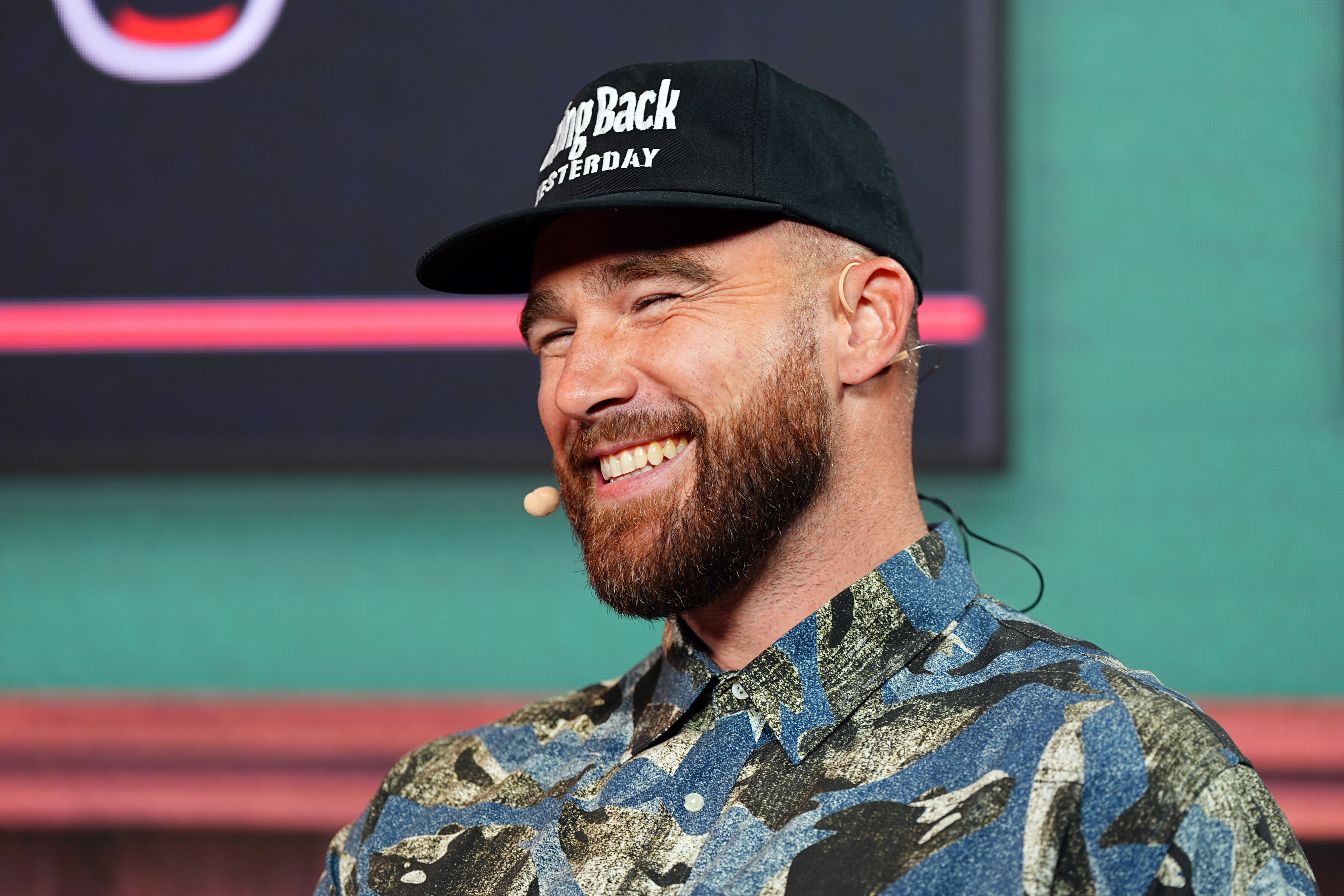 Sean Evans smiling, wearing a patterned button-up shirt and a baseball cap with the text &quot;Step Back Yesterday.&quot;