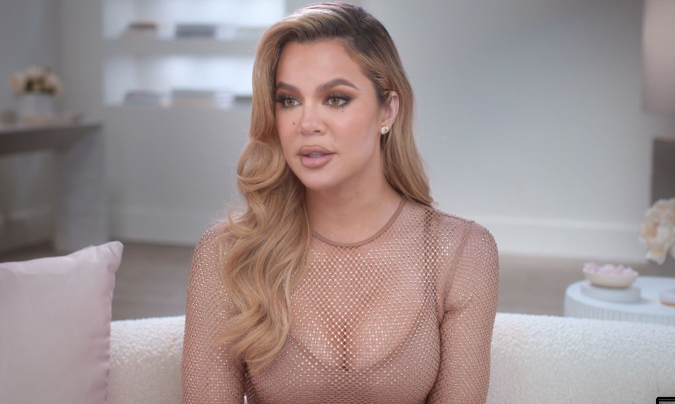 Khloé Kardashian wearing a mesh top, sits on a couch during an interview. A smaller image shows a close-up of her in the same scene