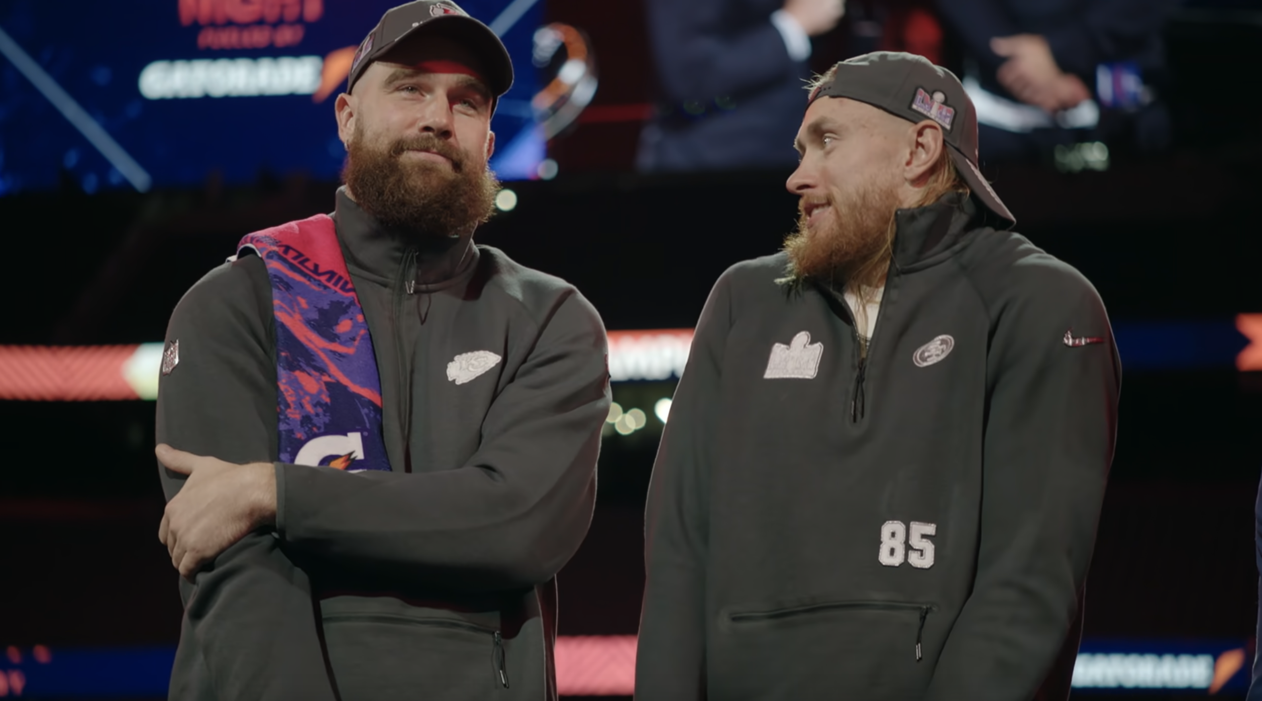 Travis Kelce and George Kittle on stage wearing casual sports hoodies and caps, smiling and interacting with each other