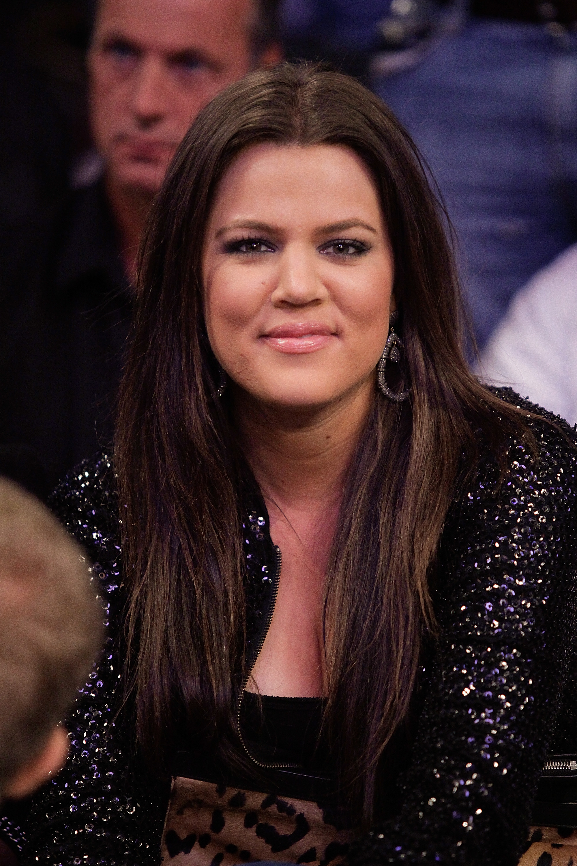 Khloé Kardashian smiling, wearing a sequined jacket and hoop earrings, with people in the background