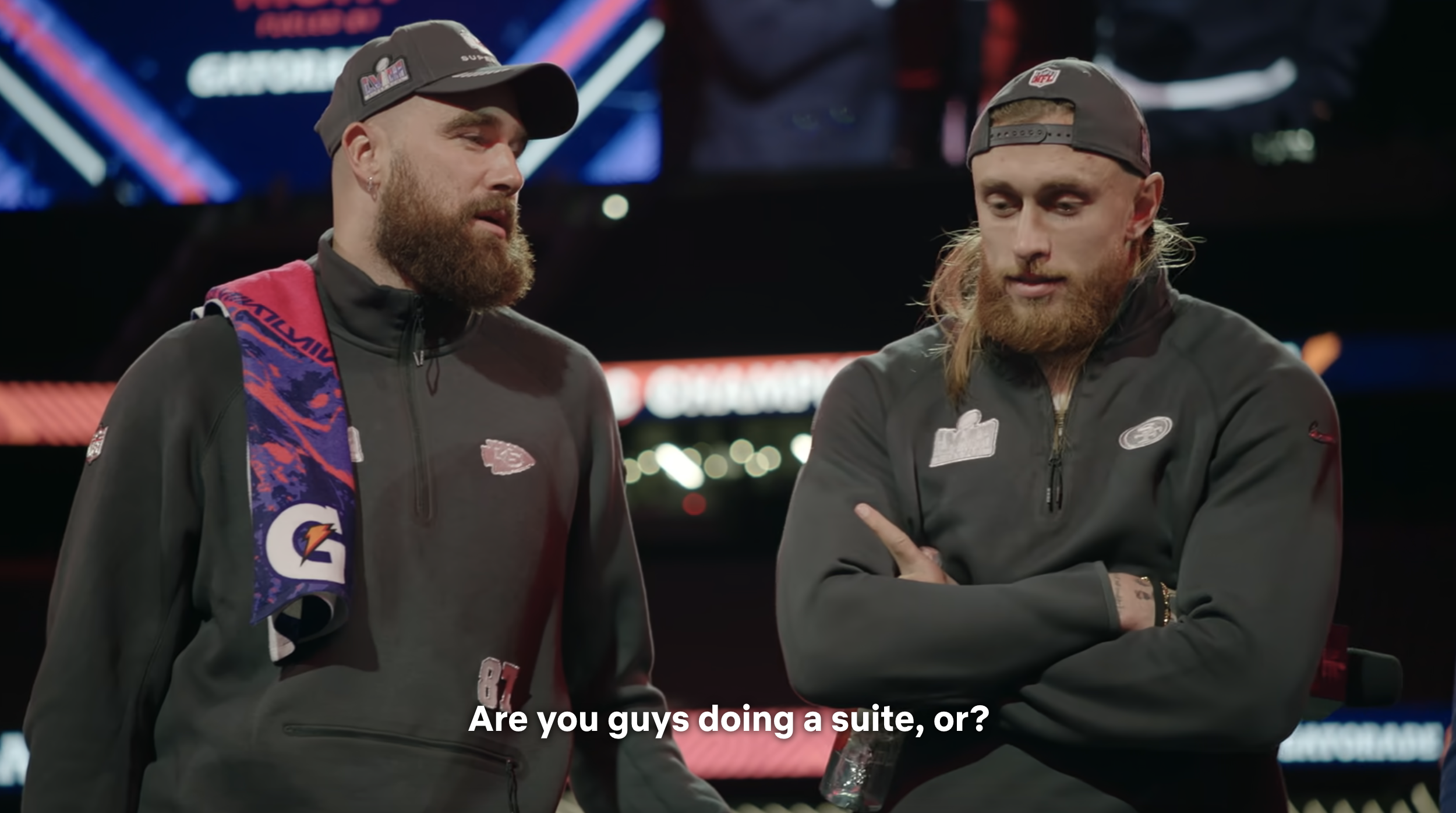 Travis Kelce and George Kittle at a media event, both wearing sports merchandise jackets. Caption: &quot;Are you guys doing a suite, or?&quot;