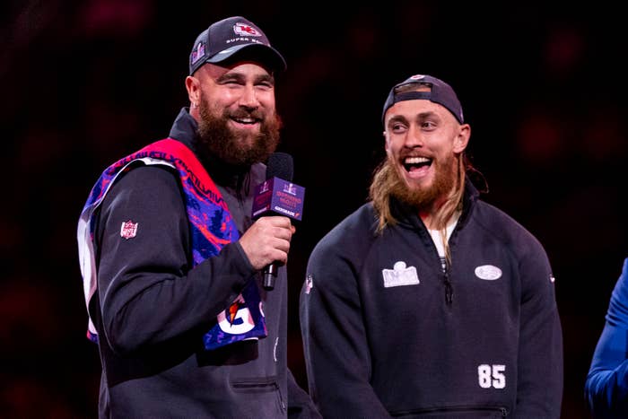 Travis Kelce (left) and George Kittle (right) are talking and laughing on stage. Both are wearing sports attire with team logos. Travis holds a microphone