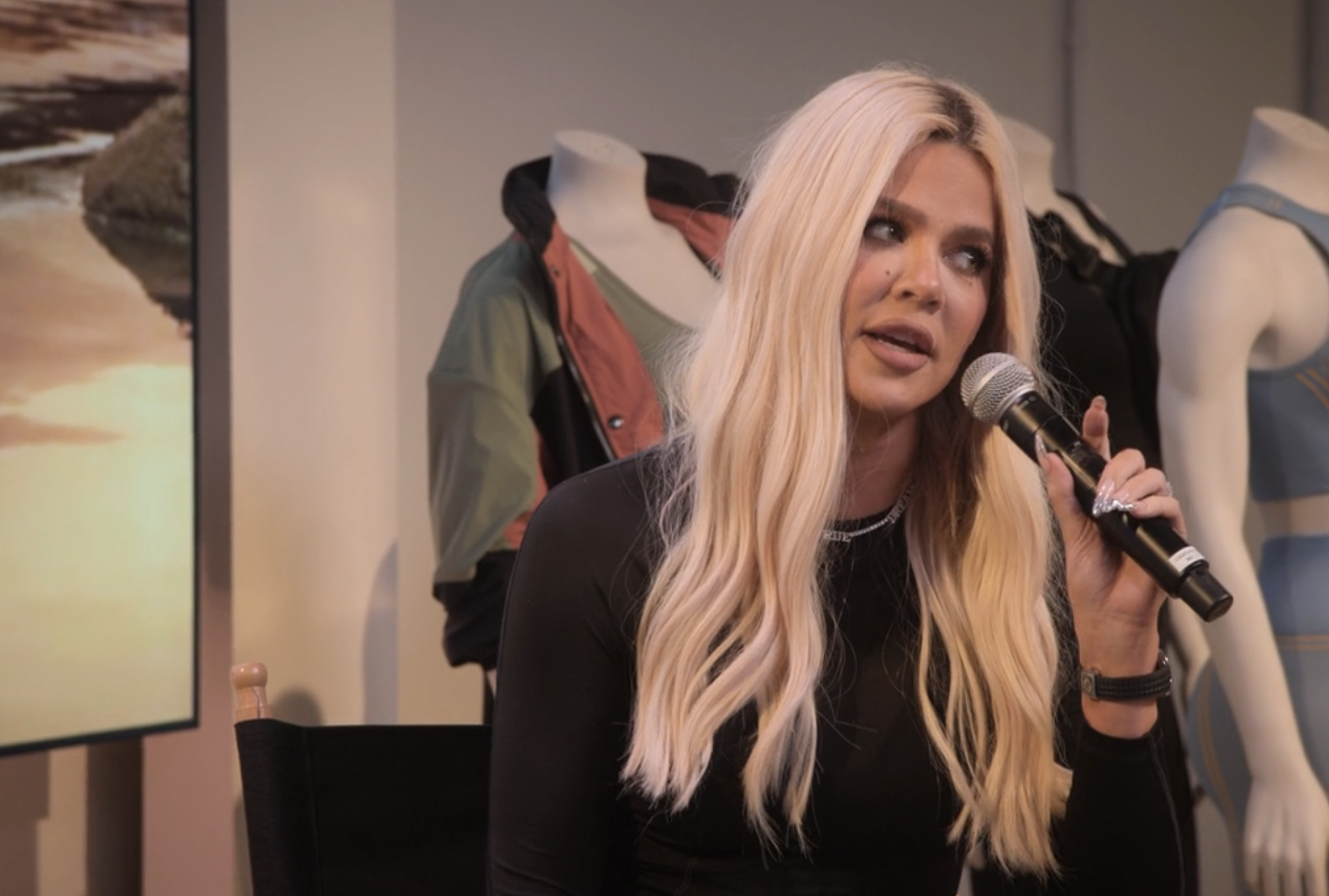 Khloé Kardashian is sitting and speaking into a microphone. Mannequins in athletic wear are behind her. A large photo print is visible to the left