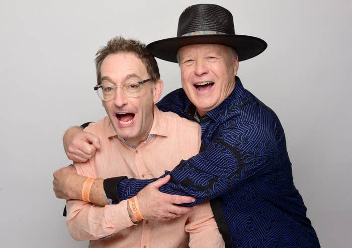 Two men happily embrace while laughing, one in a light shirt and glasses, the other in a patterned shirt and wide-brimmed hat