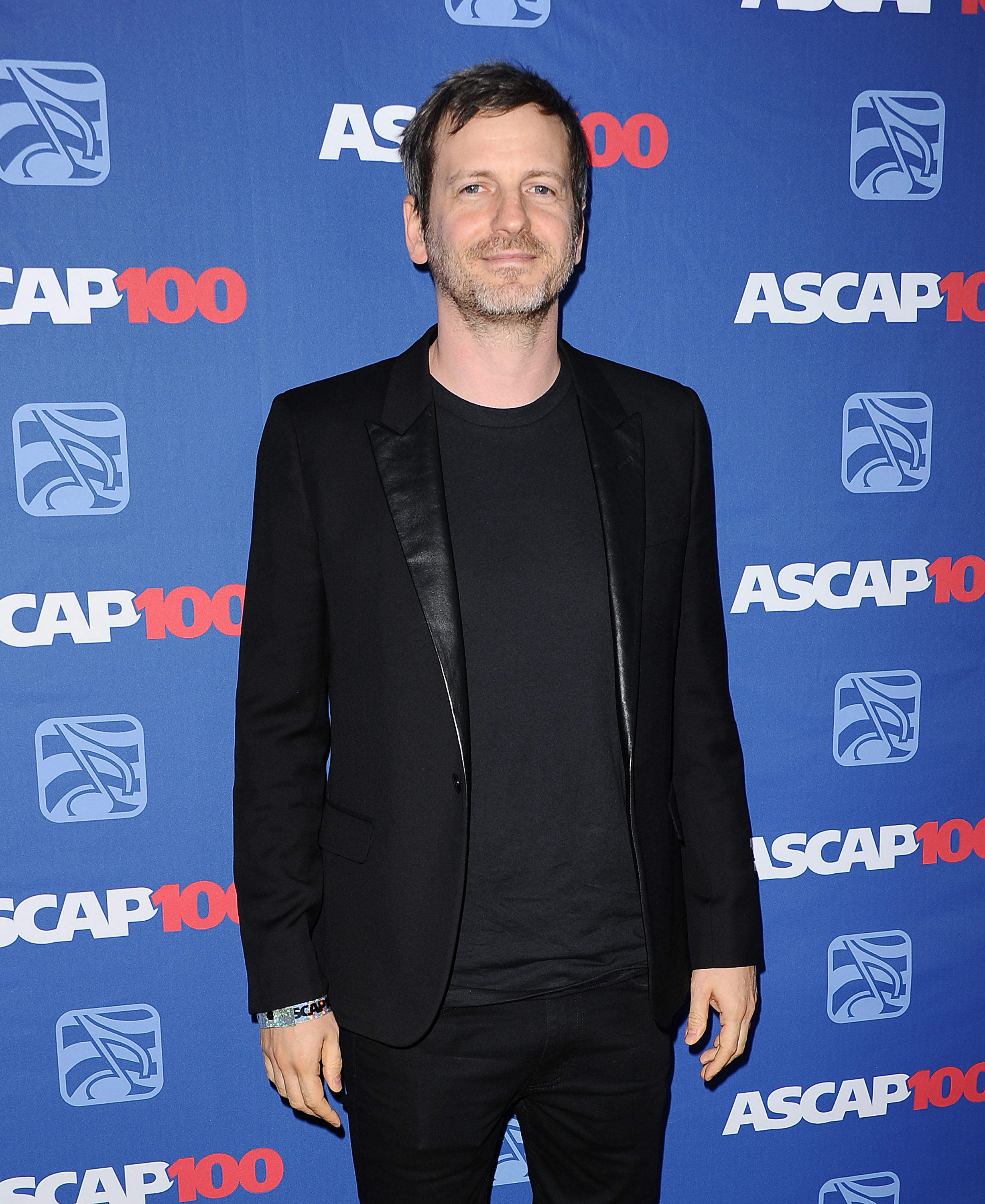 Dr. Luke at an ASCAP event, wearing a black suit with a t-shirt underneath, standing in front of a blue backdrop with ASCAP 100 logos