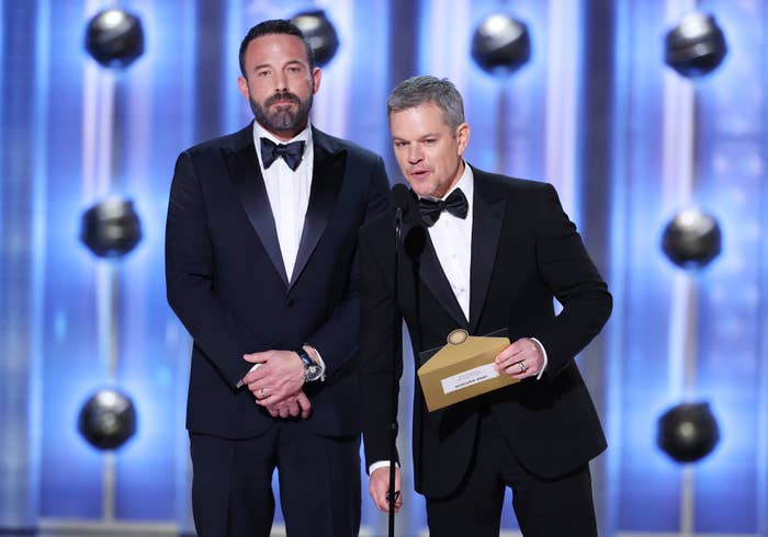Ben Affleck and Matt Damon on stage at an event, both dressed in suits with bow ties. Matt Damon is holding an envelope and speaking into a microphone