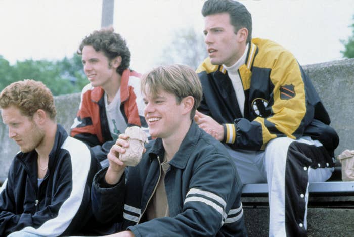 Matt Damon, Ben Affleck, and two others sit on bleachers. Damon holds a drink and smiles, while Affleck wears a jacket with a sports patch
