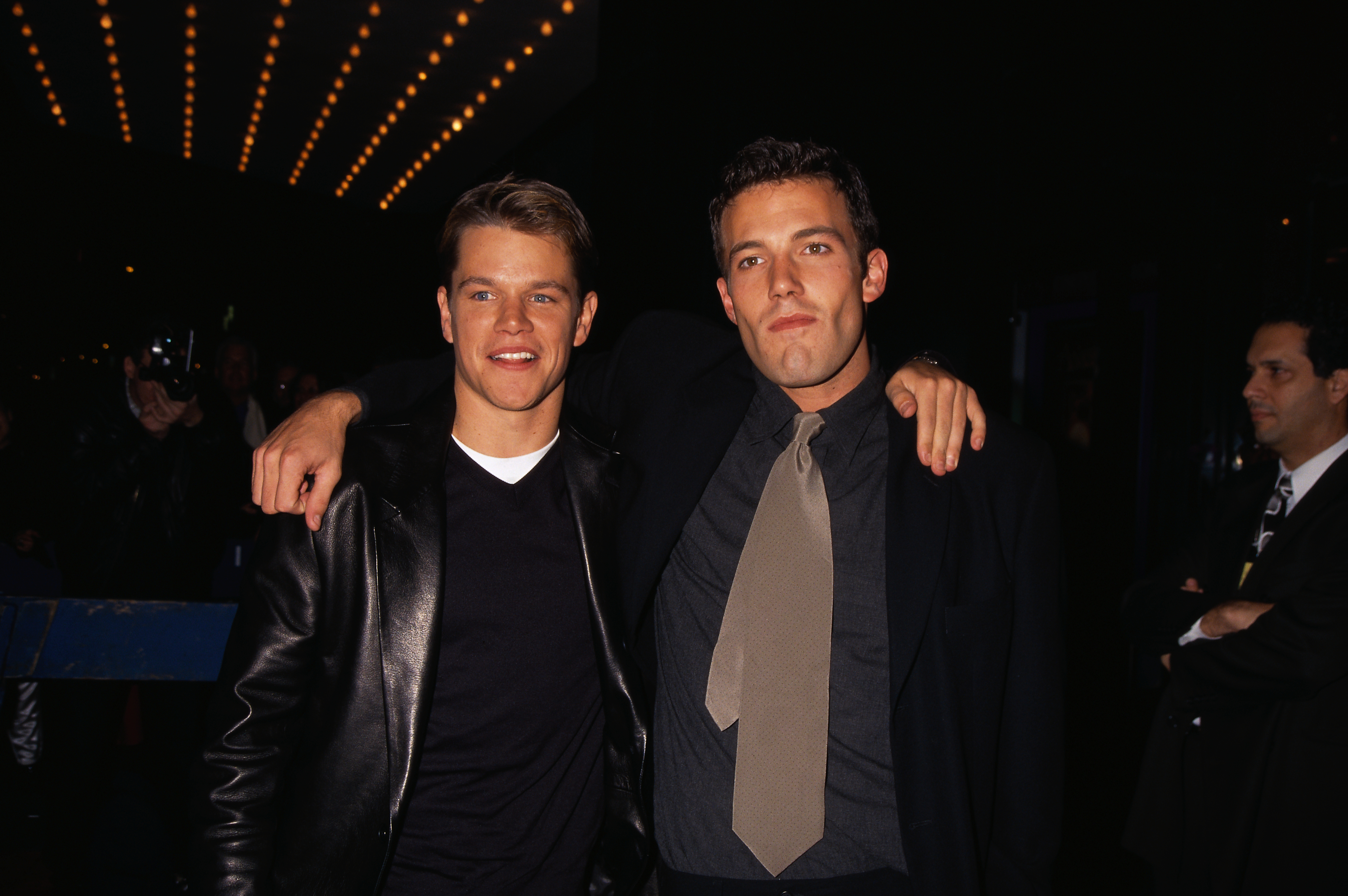 Matt Damon and Ben Affleck, in casual and formal attire respectively, pose with their arms around each other at a nighttime event