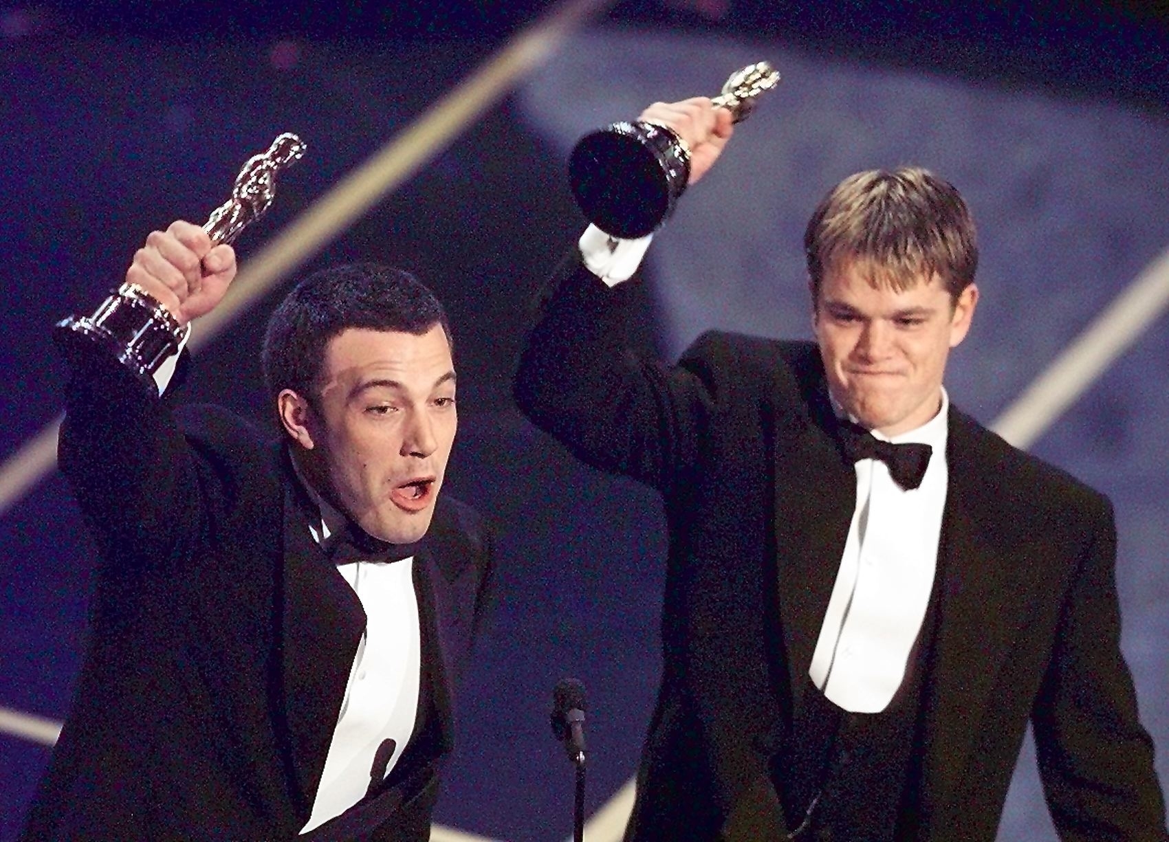 Ben Affleck and Matt Damon hold their Oscars, wearing black tuxedos, on stage at an awards ceremony