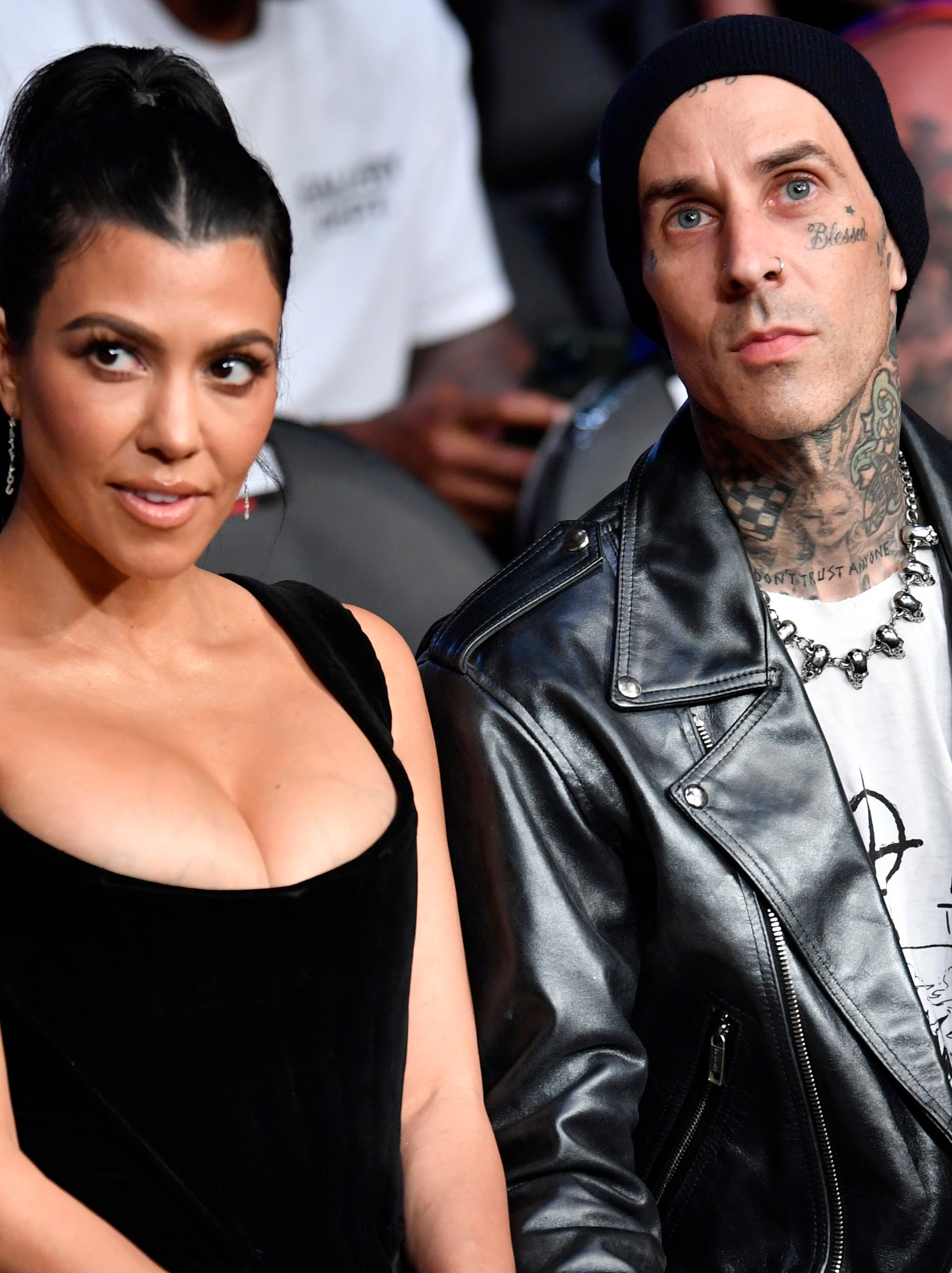 Kourtney Kardashian and Travis Barker seated at an event. Kourtney wears a low-cut dress, while Travis sports a leather jacket and beanie, showcasing his tattoos