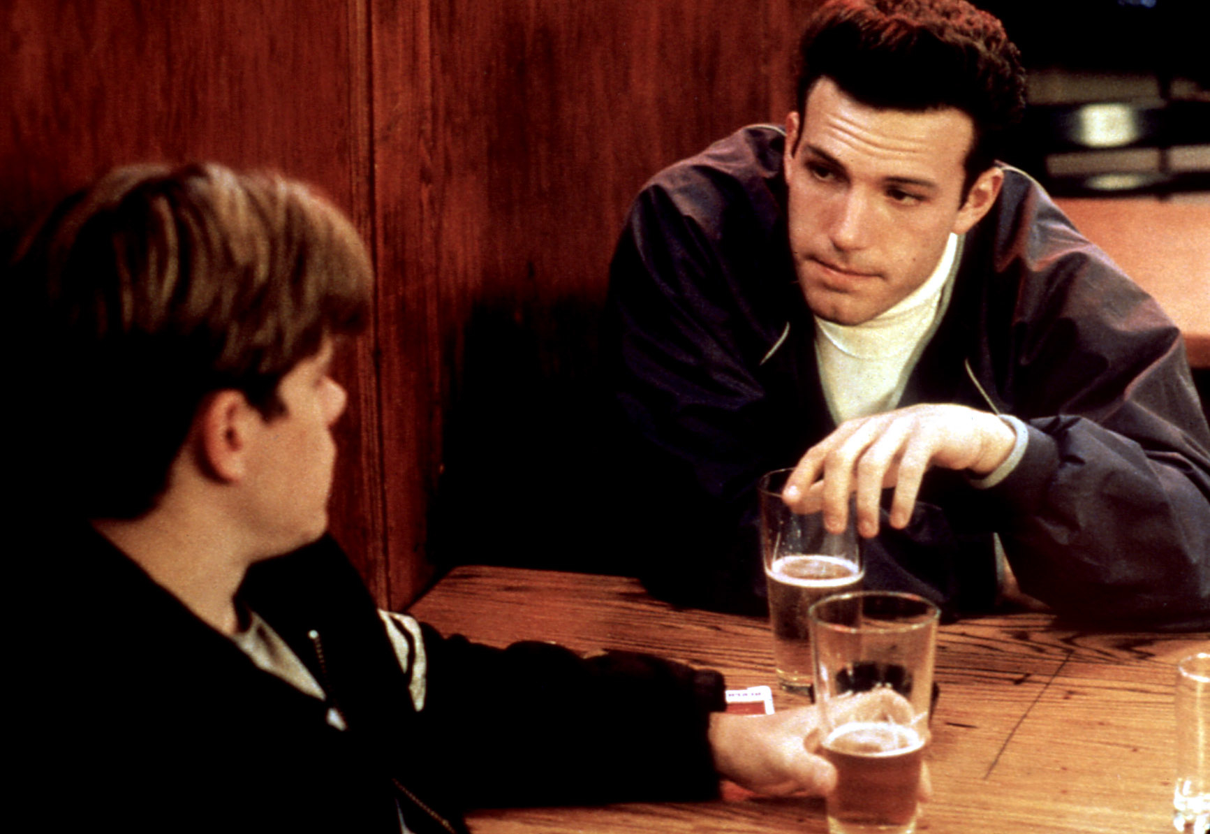 Matt Damon and Ben Affleck are seated at a wooden table, engaging in conversation over drinks in a casual setting