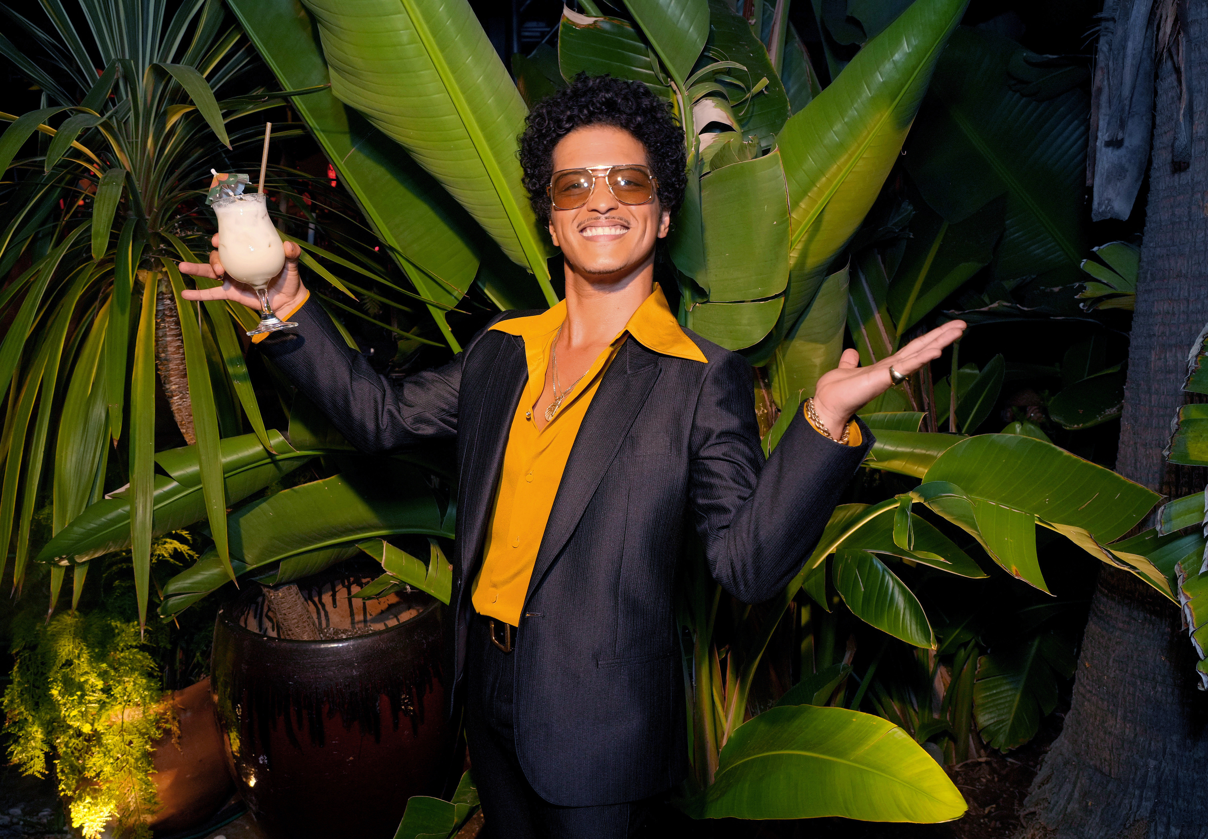 Bruno Mars is smiling, wearing a yellow shirt and black suit, holding a drink, arms outstretched, in a tropical setting