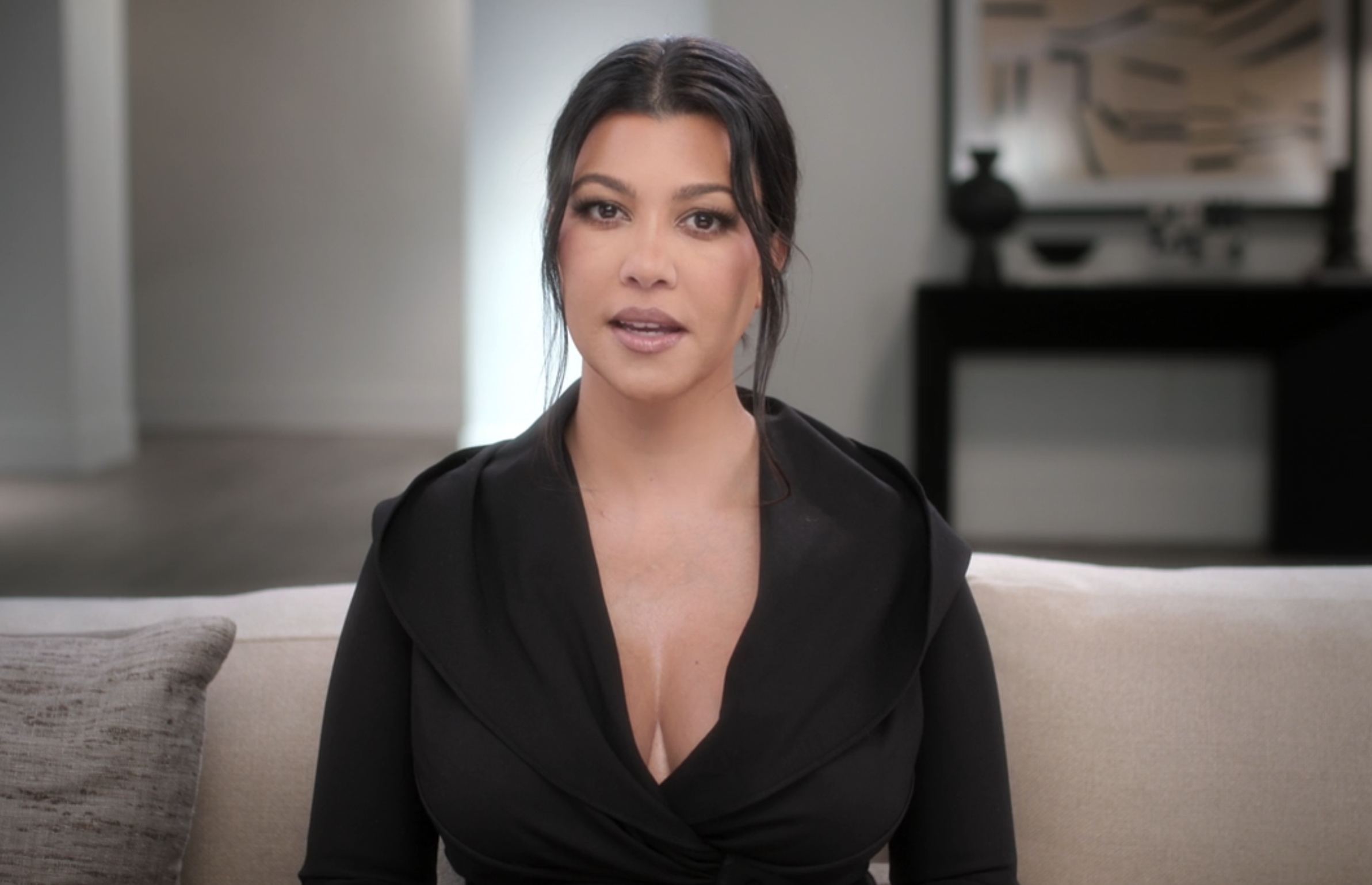 Kourtney Kardashian sits on a couch in a modern living room, wearing a stylish black v-neck top. She is facing the camera, delivering a message