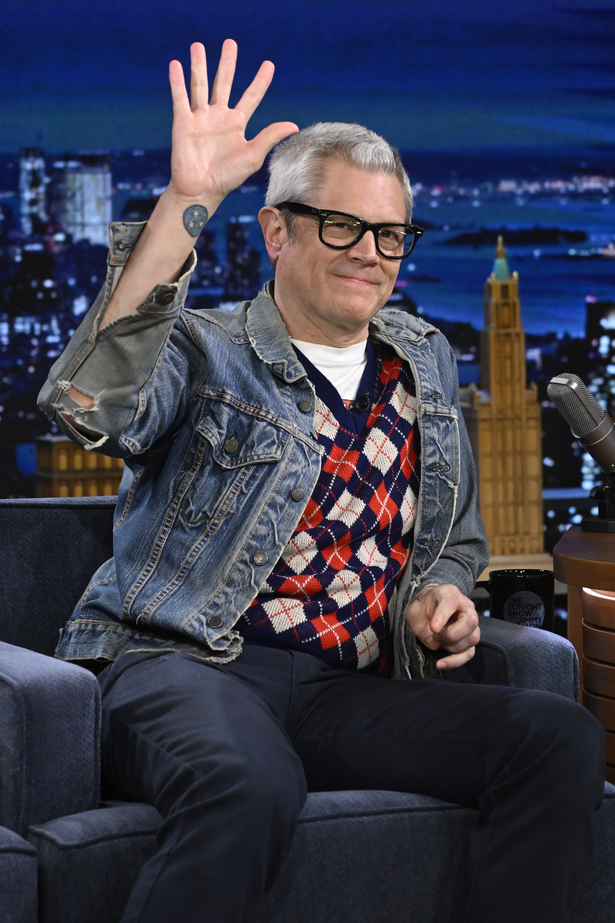 Johnny Knoxville, dressed in a denim jacket, patterned sweater, and glasses, waves while seated on a talk show set