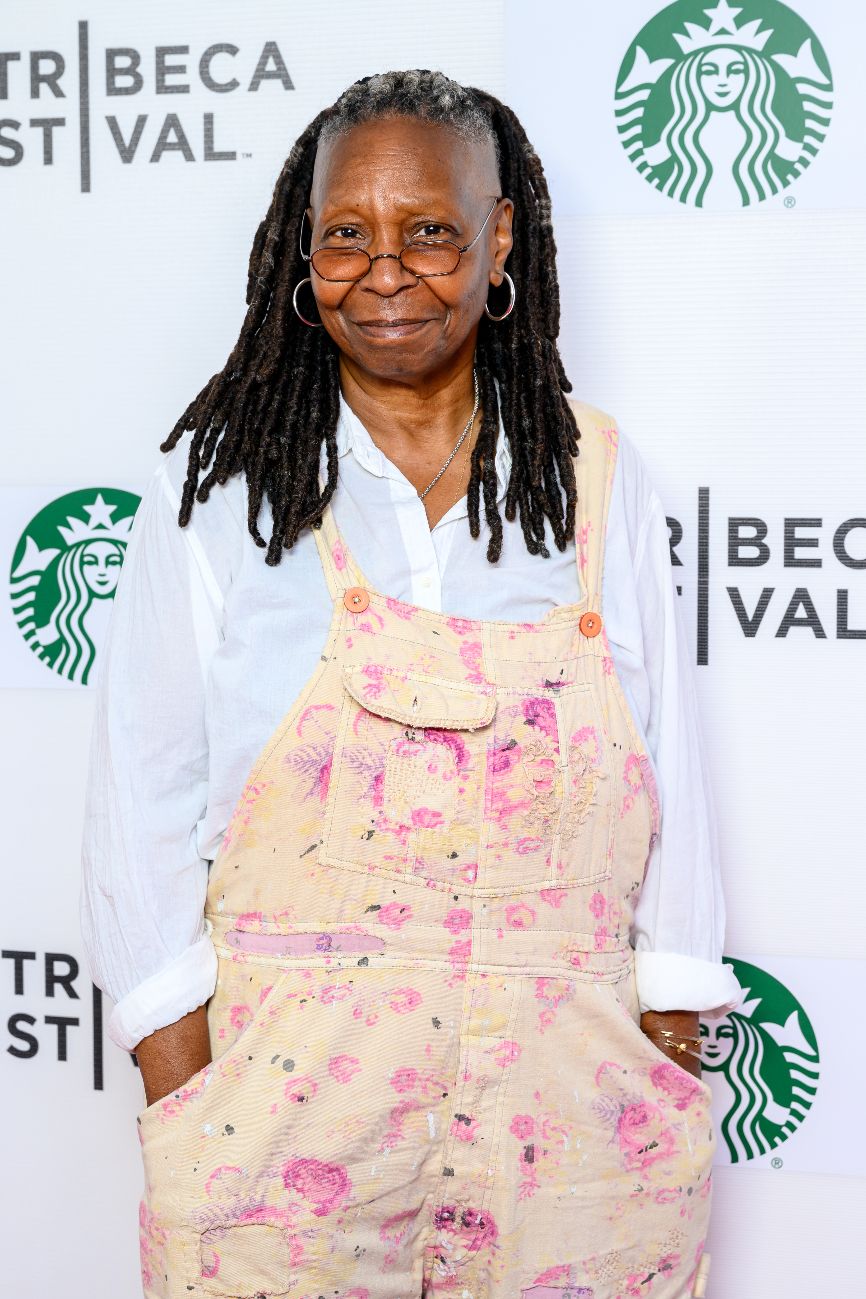 Whoopi Goldberg at Tribeca Festival event in paint-splattered overalls and a white shirt, with dreadlocks and round glasses, standing in front of a Starbucks logo backdrop